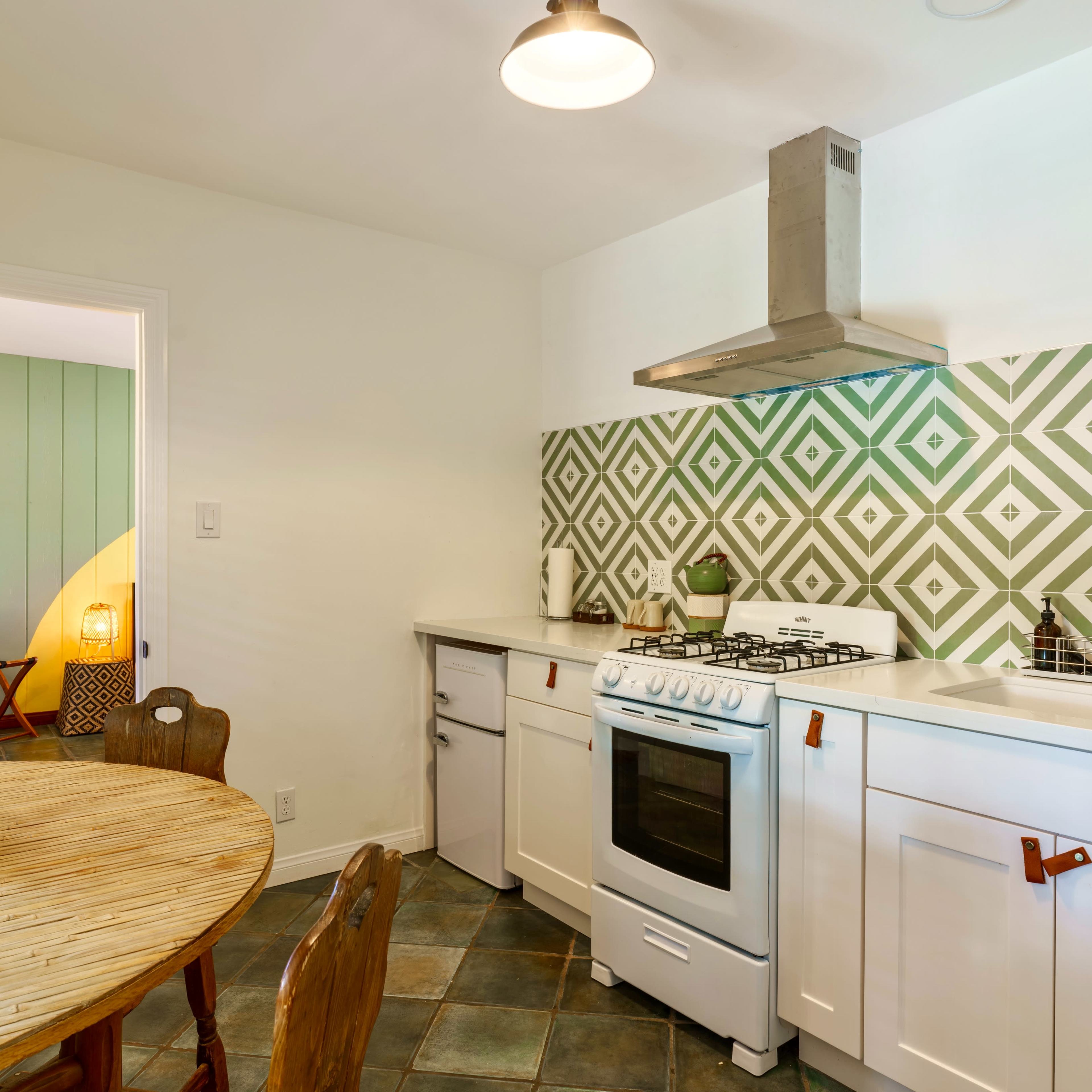A kitchen features a gas stove, white cabinetry, and a patterned green backsplash, alongside a wooden dining table with chairs.
