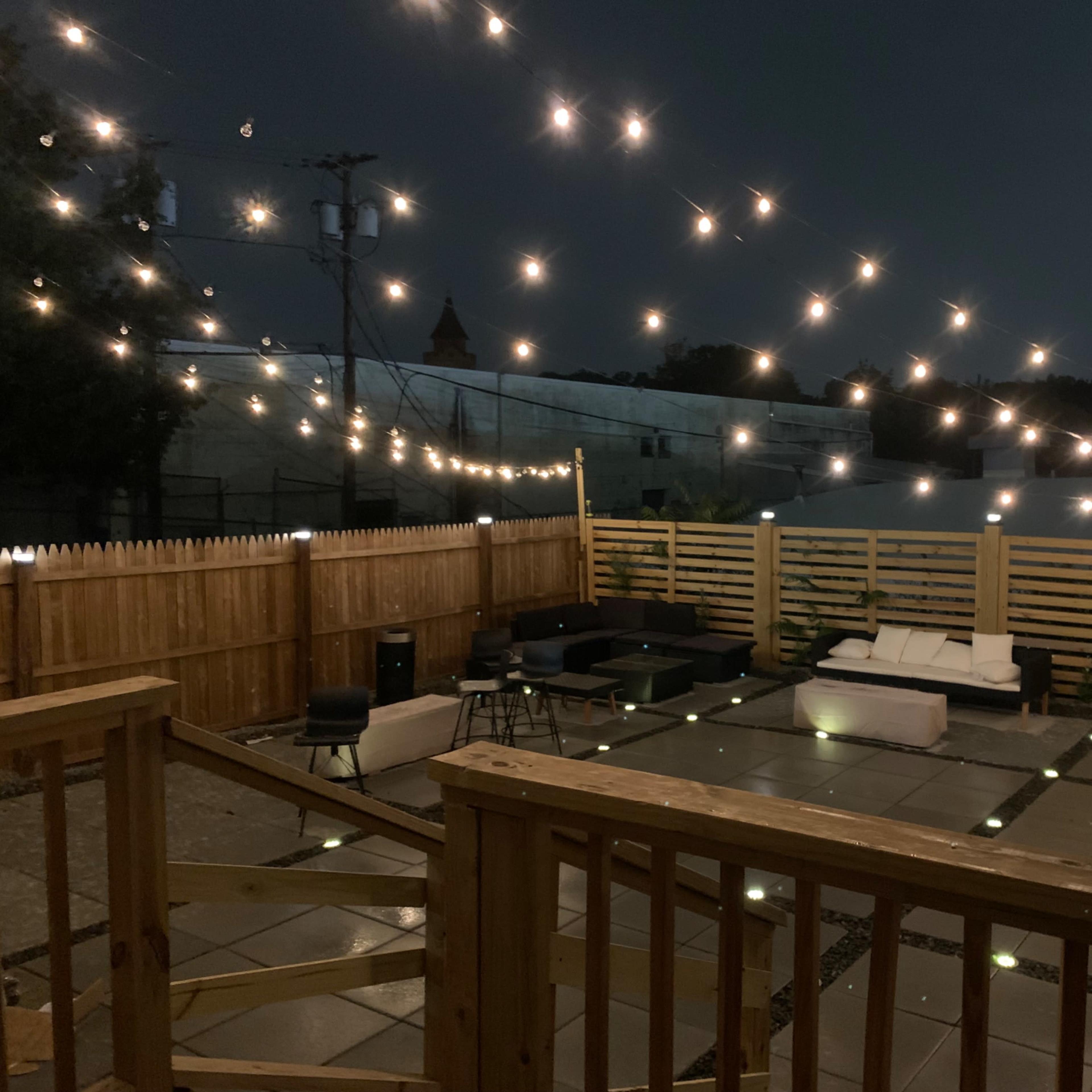 A patio area features a stone floor illuminated by string lights overhead, surrounded by wooden fencing and various seating arrangements.