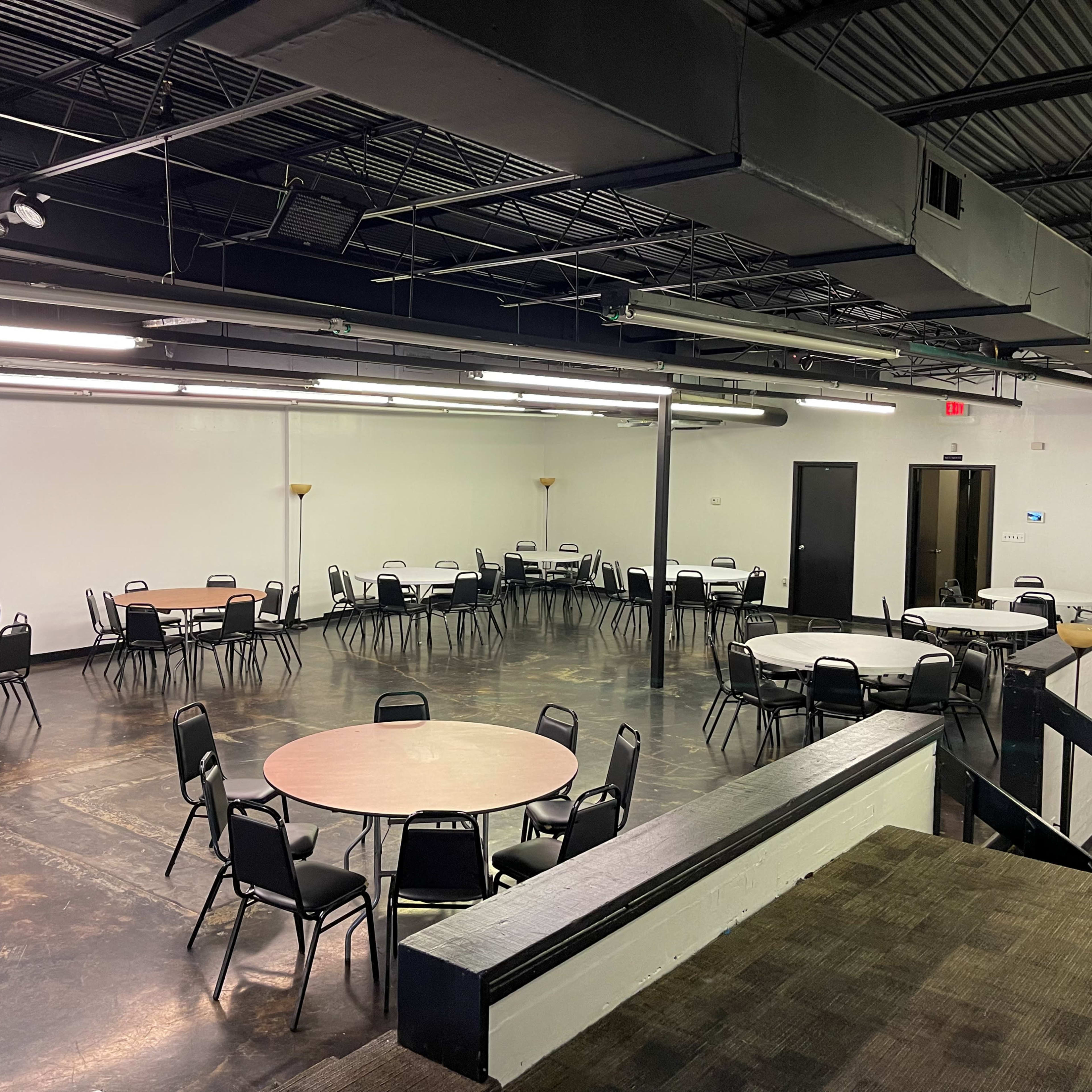A spacious, empty hall with several round tables and chairs arranged throughout.