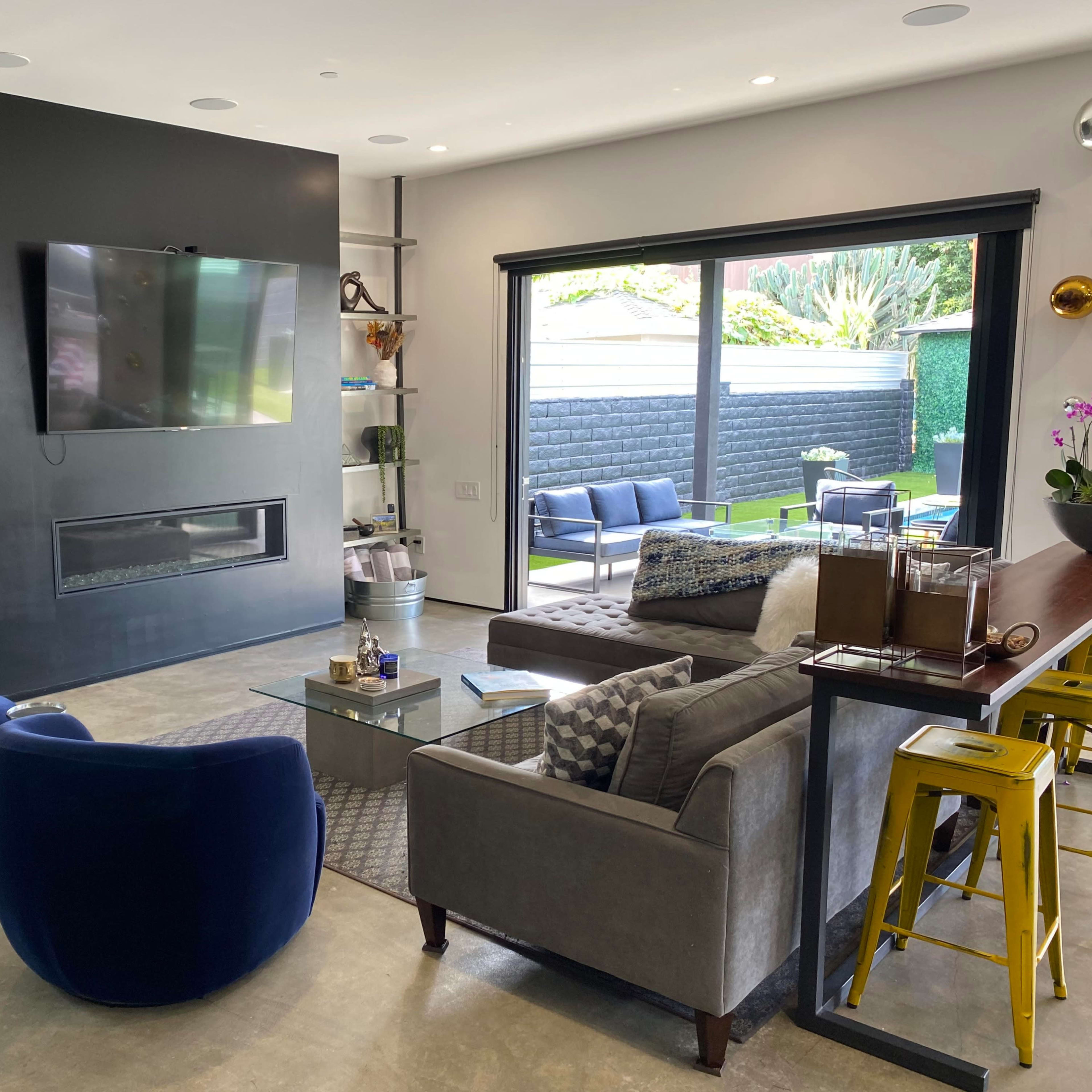 The modern living room features a television mounted on a black wall, a gray sofa, blue accent chairs, and large sliding glass doors leading to a patio area.