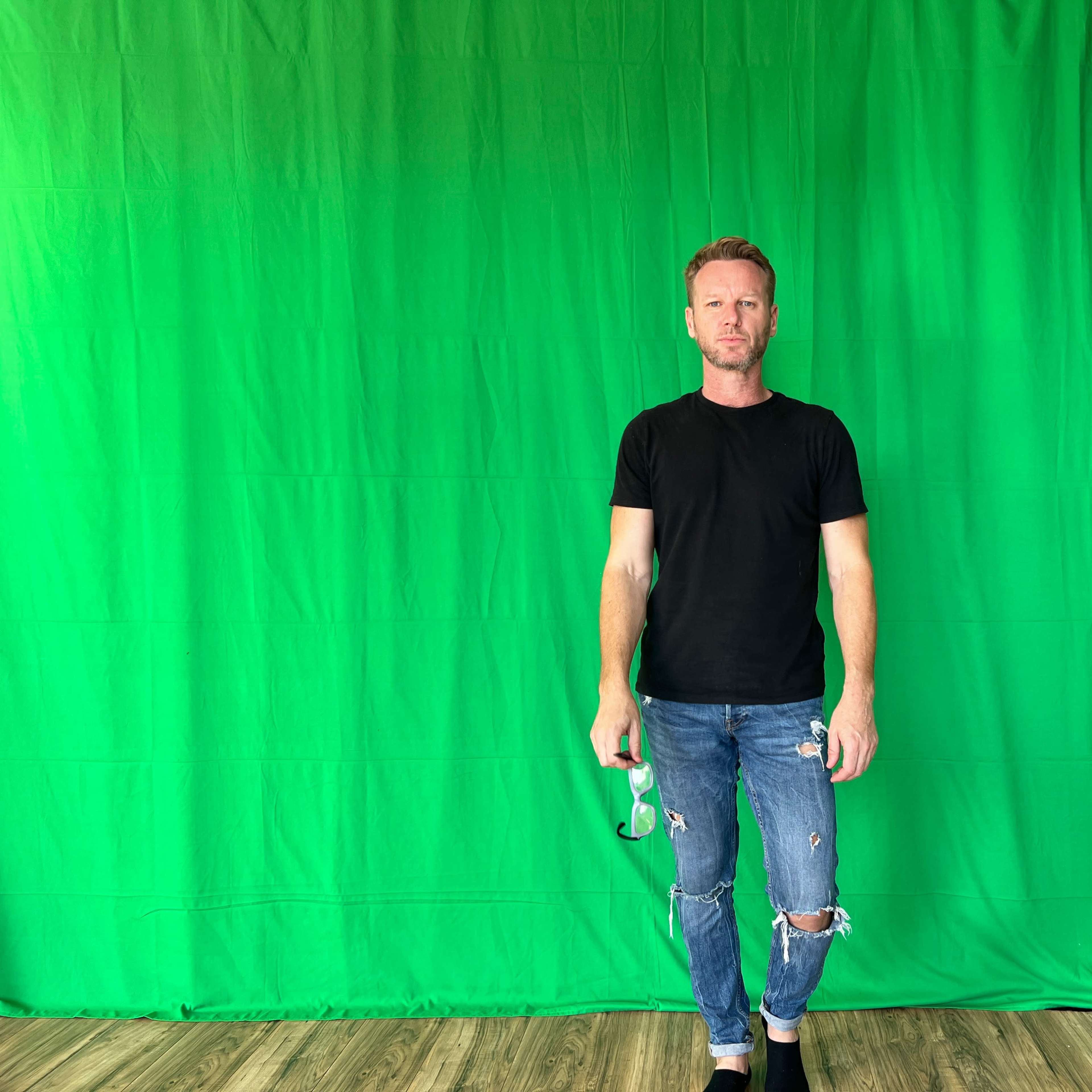 A man in a black t-shirt and ripped jeans stands on a wooden floor in front of a green backdrop.