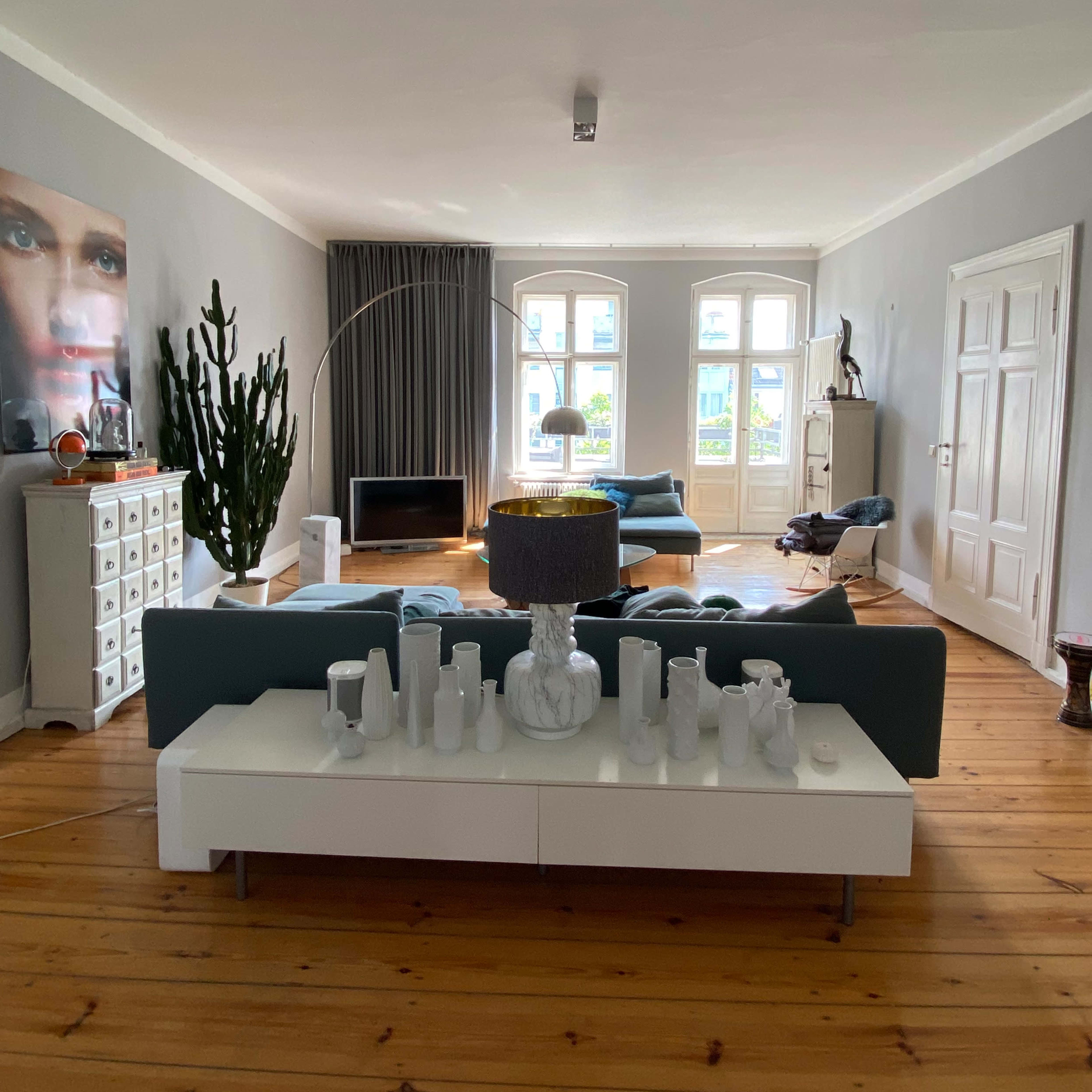 A spacious living room featuring green sofas, a white coffee table adorned with various vases, large windows with curtains, and a tall indoor plant.