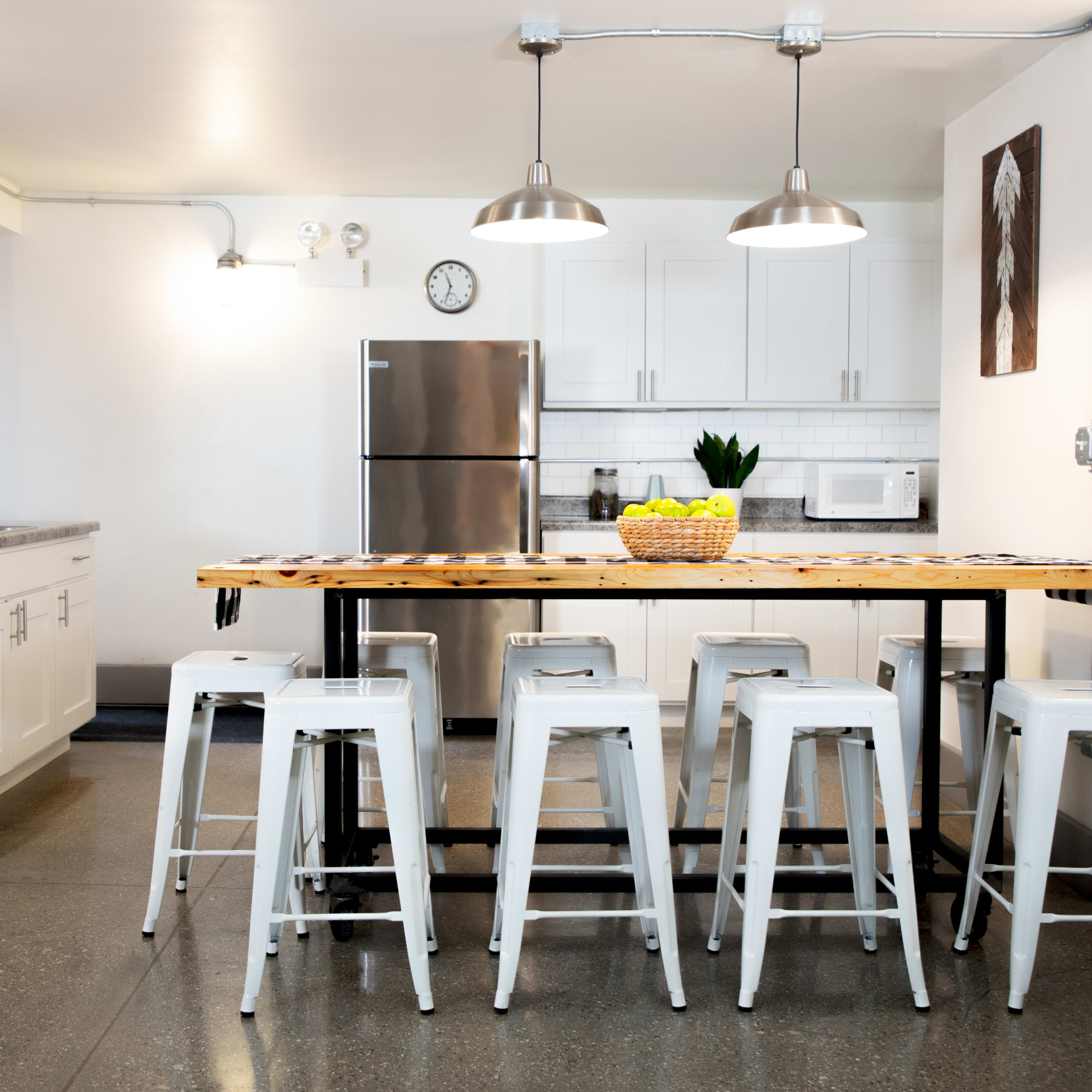 A modern kitchen features a large wooden table with metal stools, stainless steel appliances, and white cabinetry.