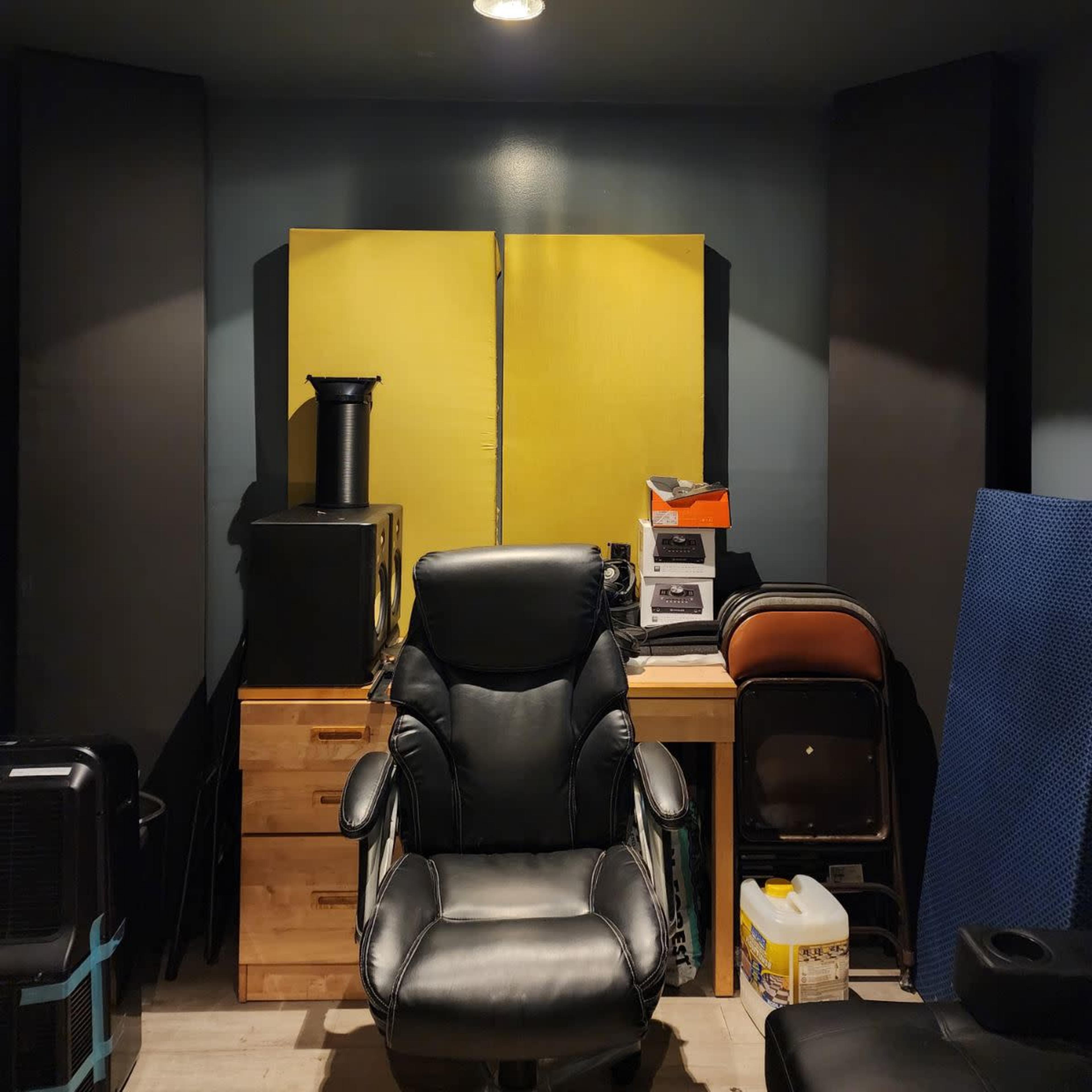 A small room with a black leather office chair, a wooden cabinet, and various audio equipment against the walls, which are partially covered with soundproofing panels.