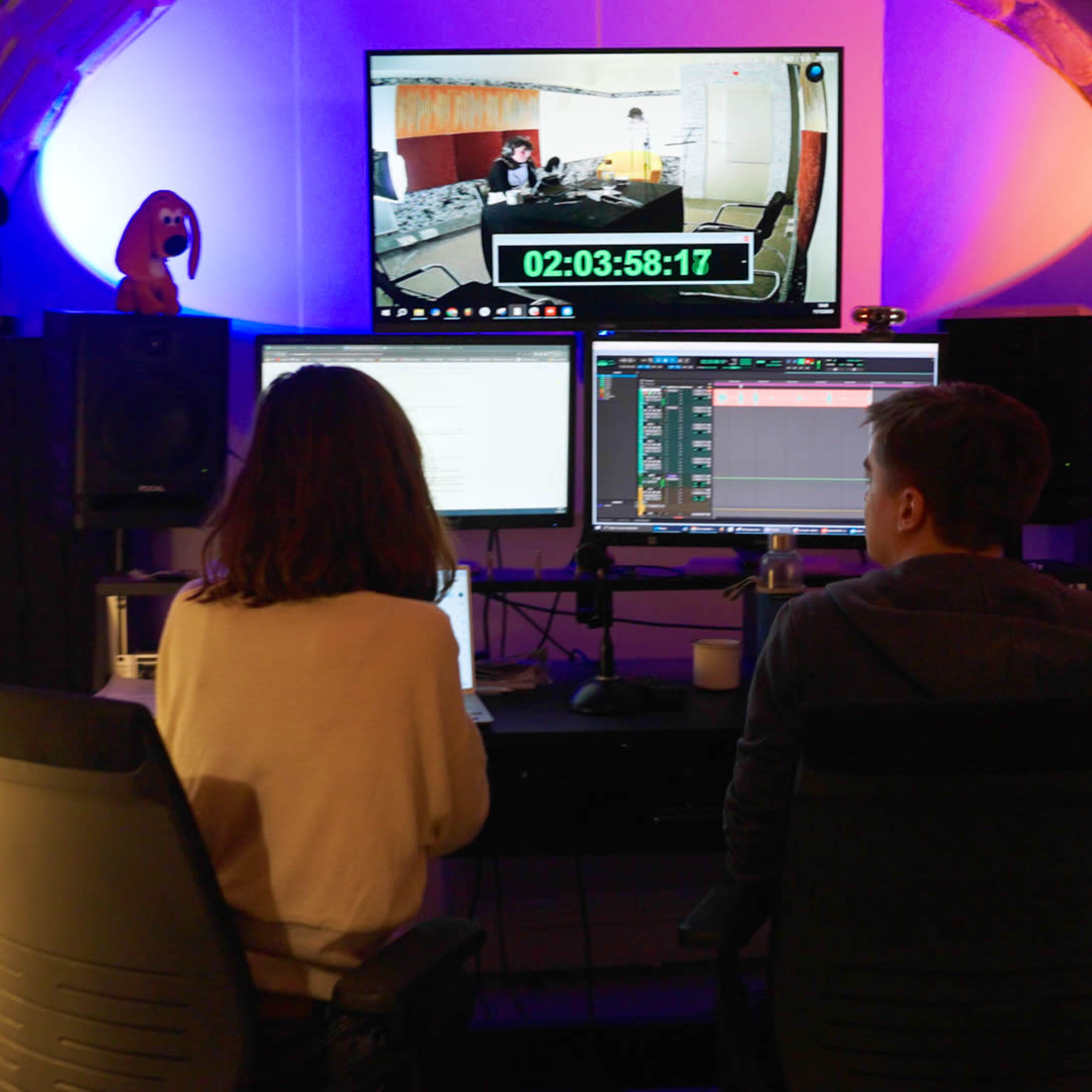 Two individuals sit at a dual-monitor workspace in a studio, facing a large screen displaying a timer and a video feed.