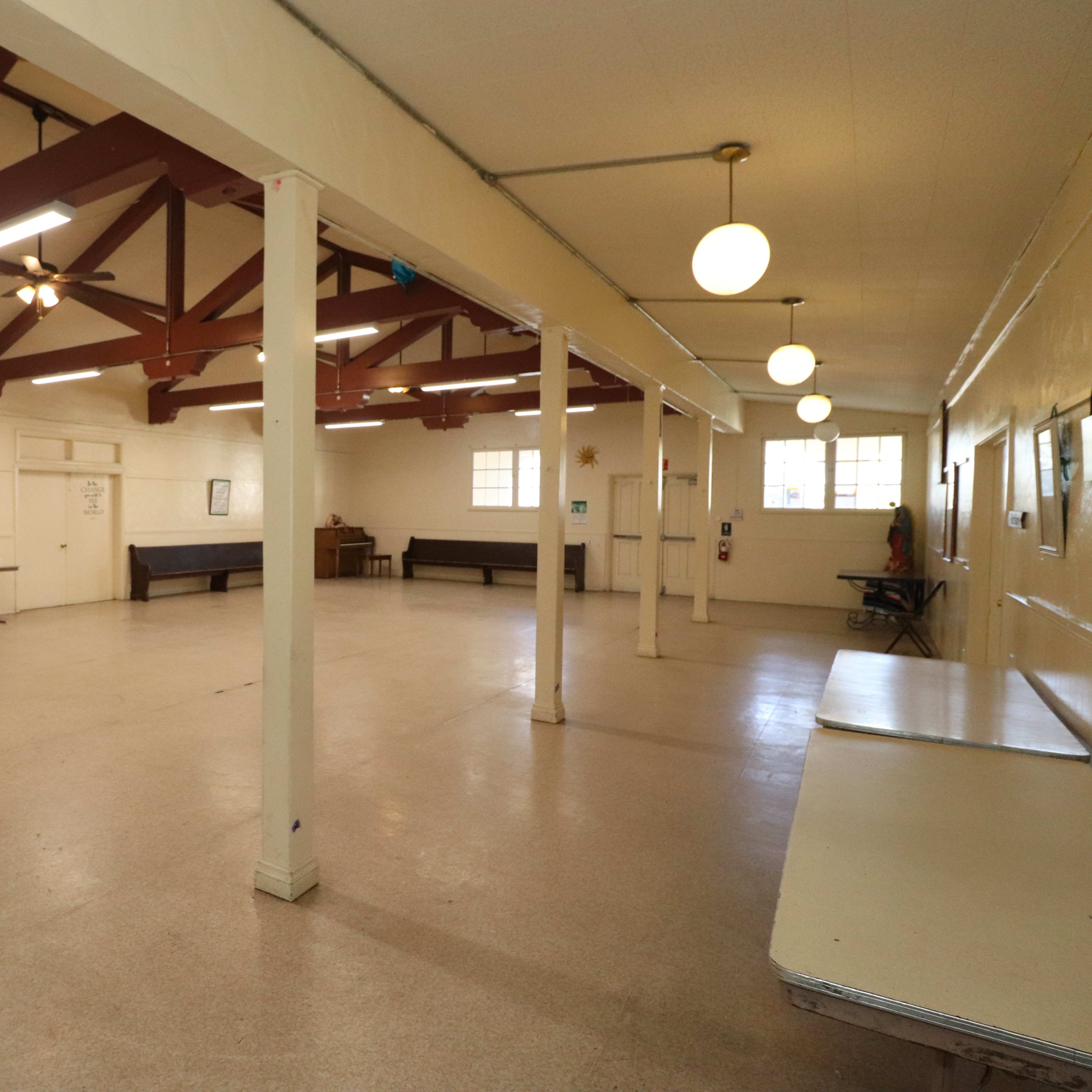 A spacious indoor room with a high ceiling, wooden beams, and several tables along the edges.