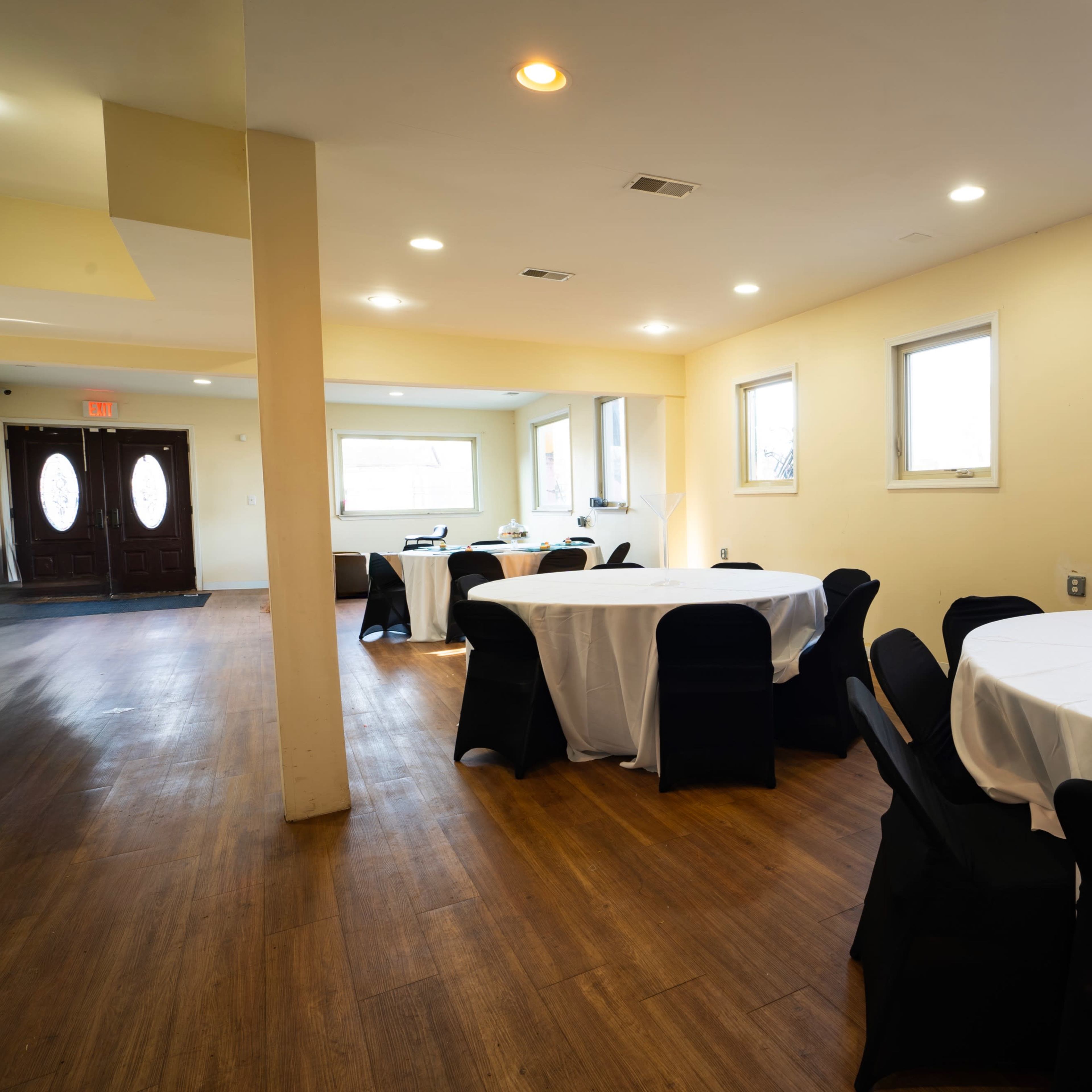 A brightly lit indoor venue featuring several round tables covered with white tablecloths and black chairs arranged along a wooden floor.