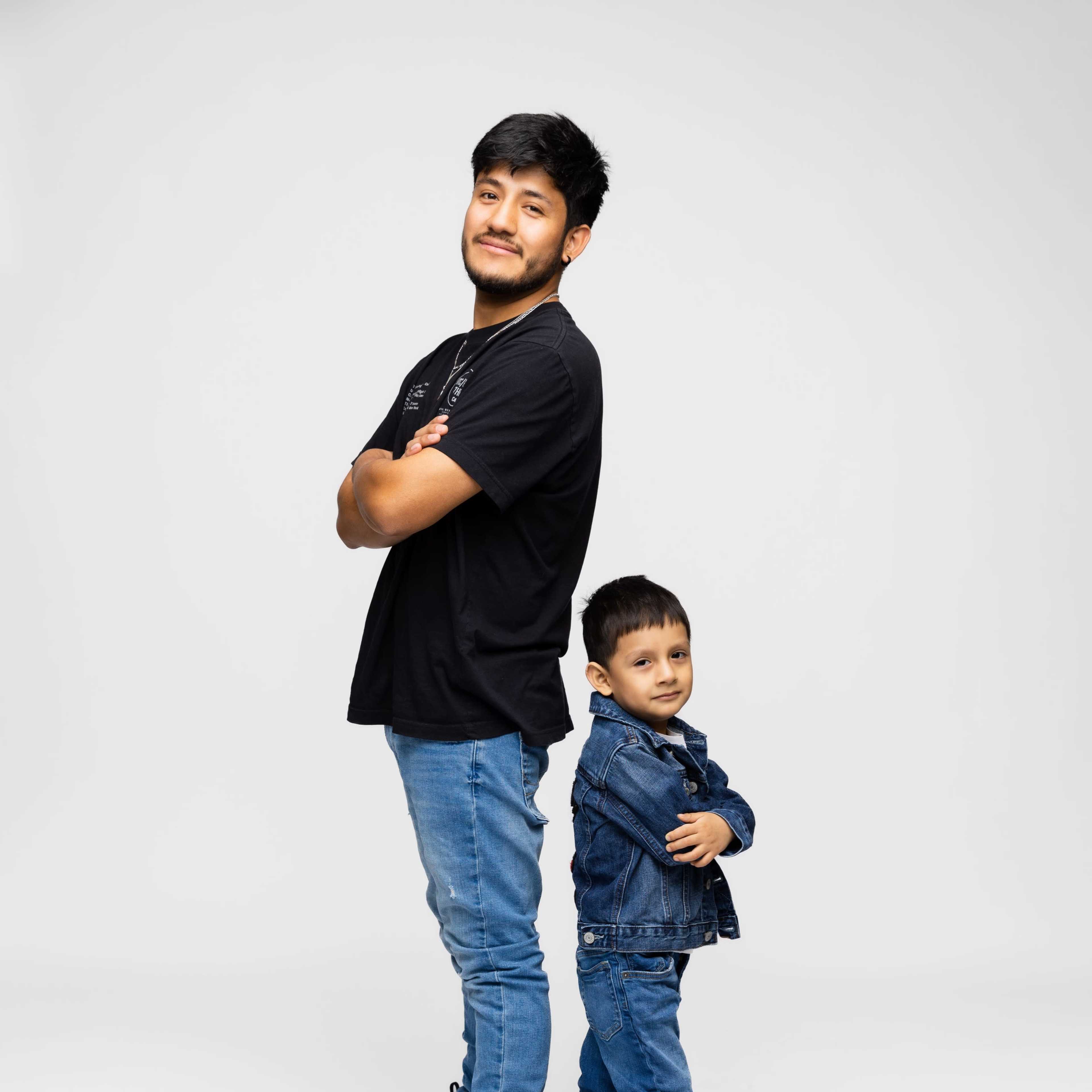 A young man stands confidently with his arms crossed, while a young boy in a denim jacket poses behind him, also with crossed arms.