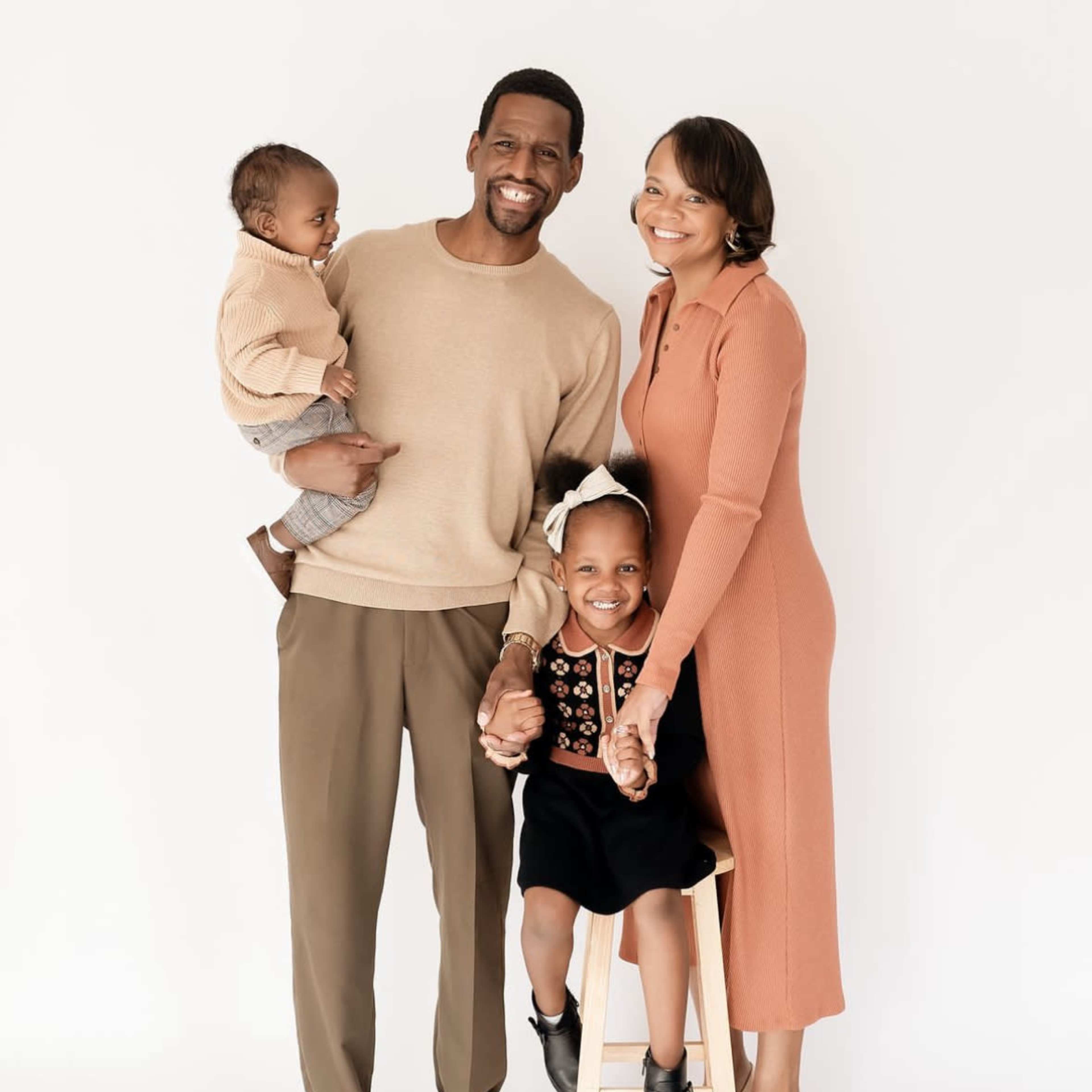 A family of four poses together, with one parent holding a toddler and the other parent holding a young child on a stool.