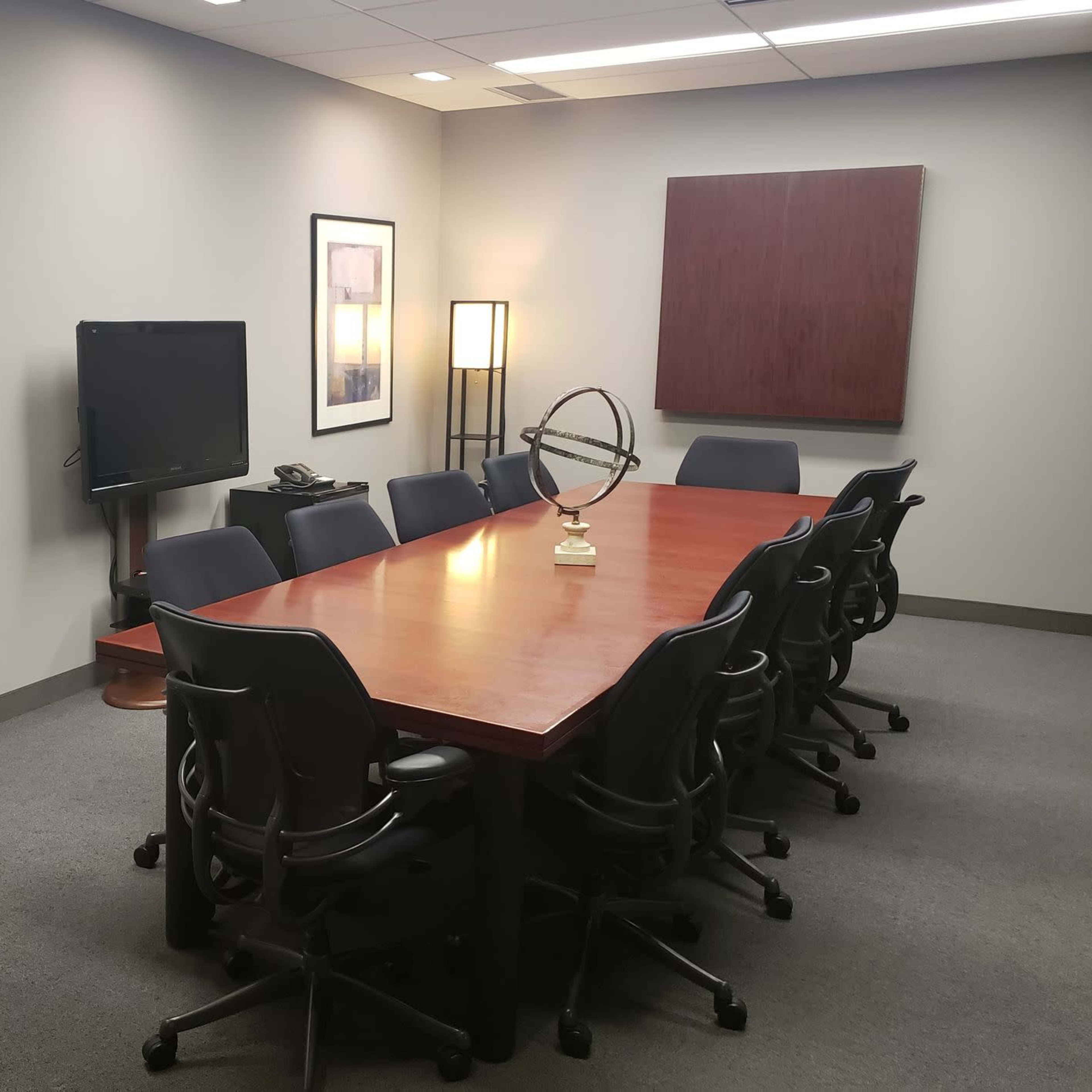 A conference room with a large wooden table surrounded by chairs, a television on the wall, and framed artwork.