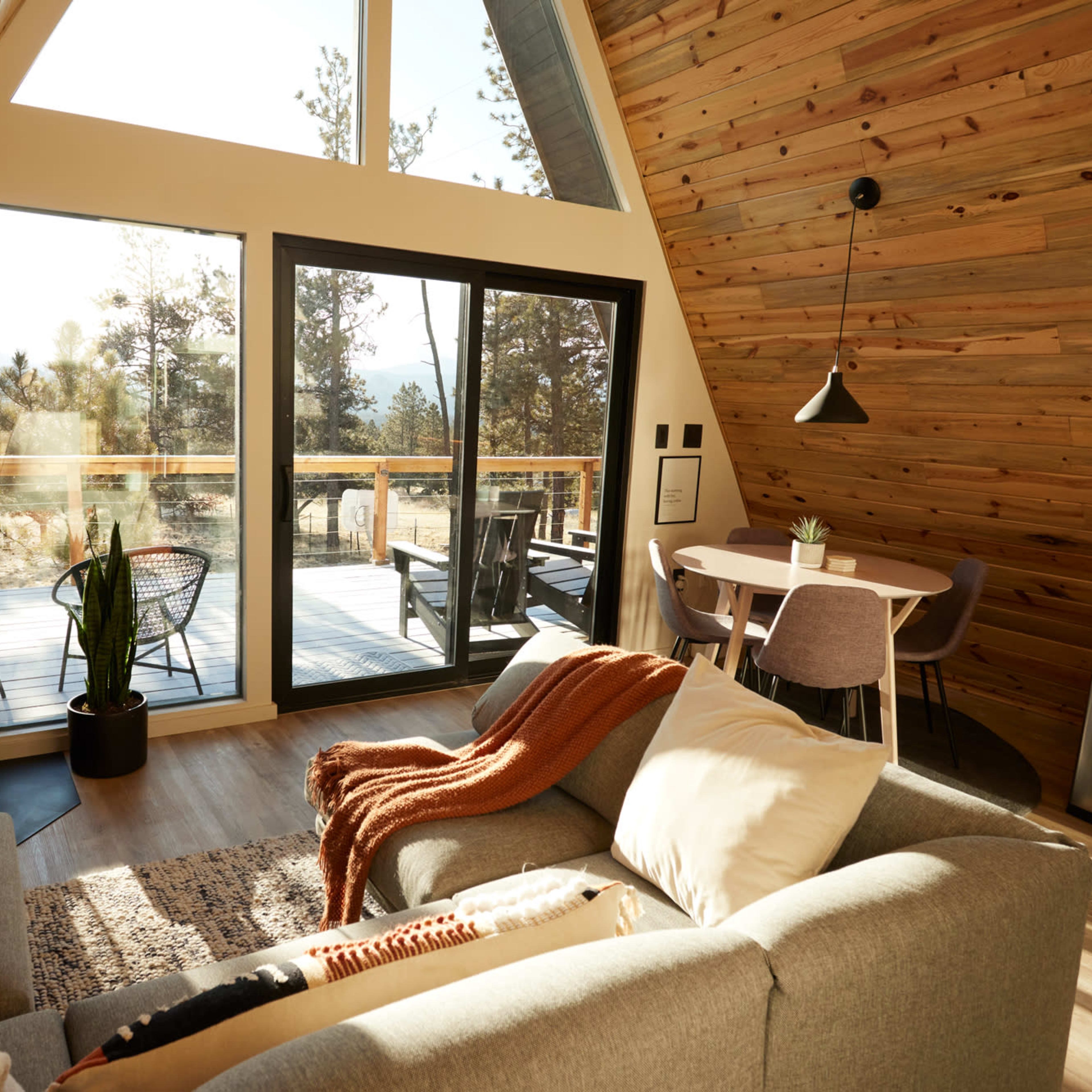 The interior of a modern A-frame cabin features a cozy seating area, large windows that overlook a wooden deck, and a dining space with minimalistic furniture.