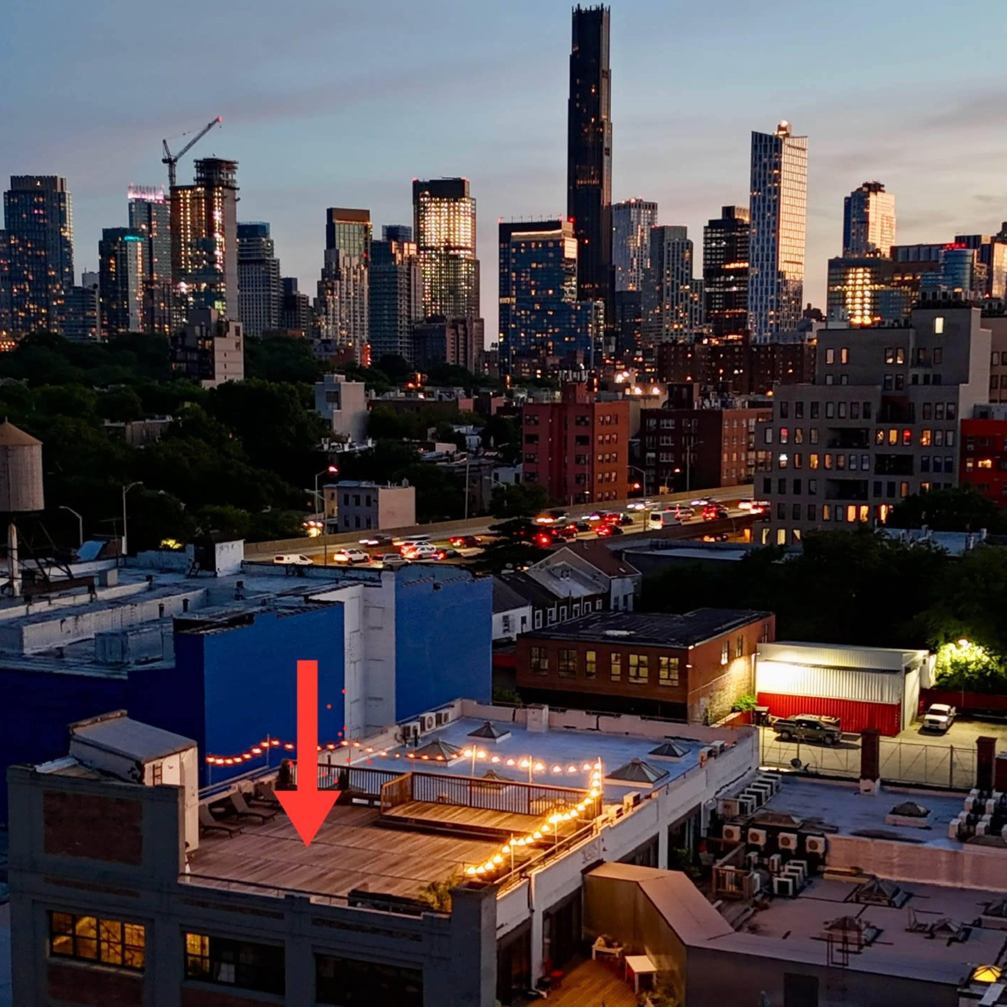 A city skyline at dusk with skyscrapers illuminated against a fading sky, and a rooftop with string lights in the foreground.