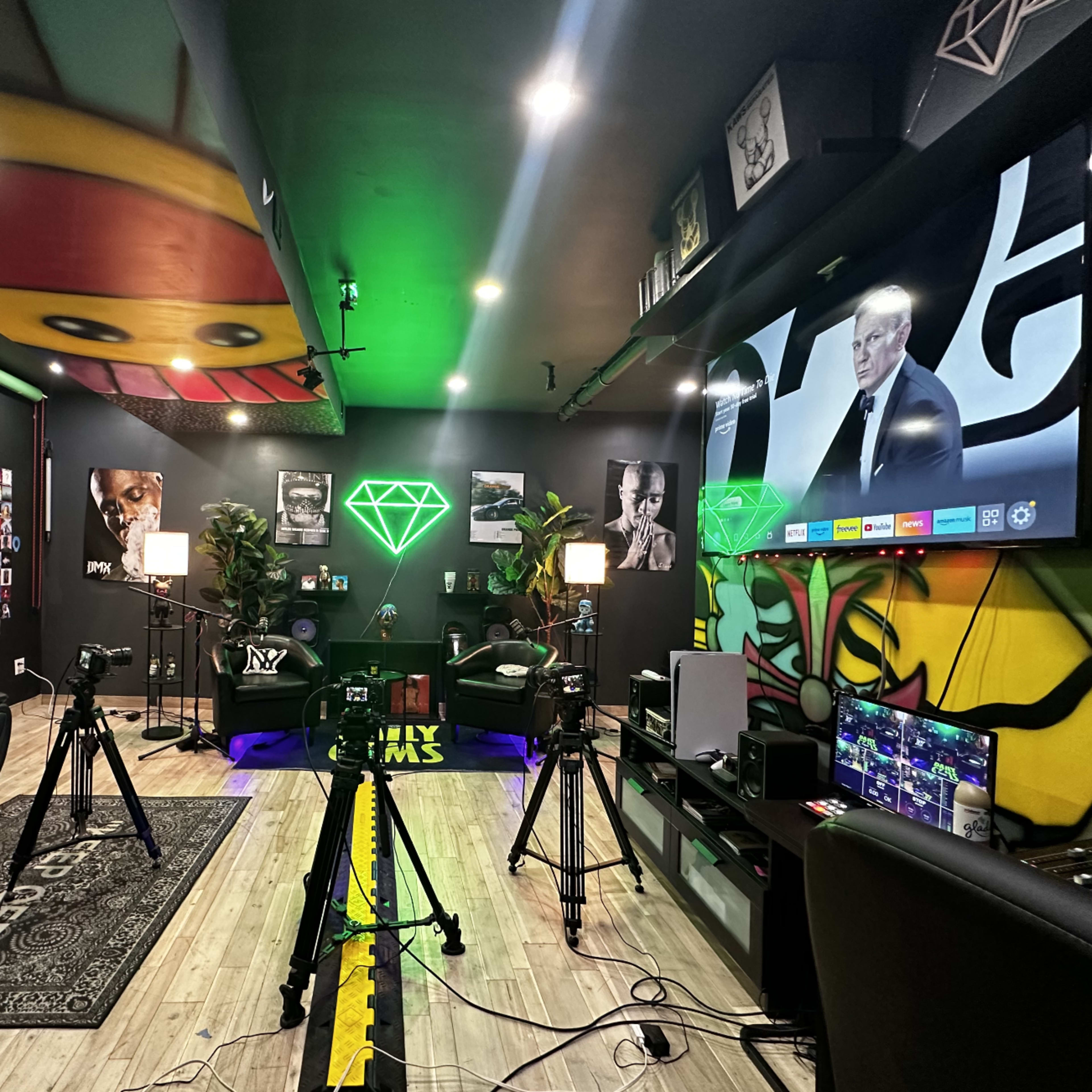 A modern multimedia studio setup with green lighting, cameras, and a large screen displaying a presentation, surrounded by vibrant wall art and photographs.