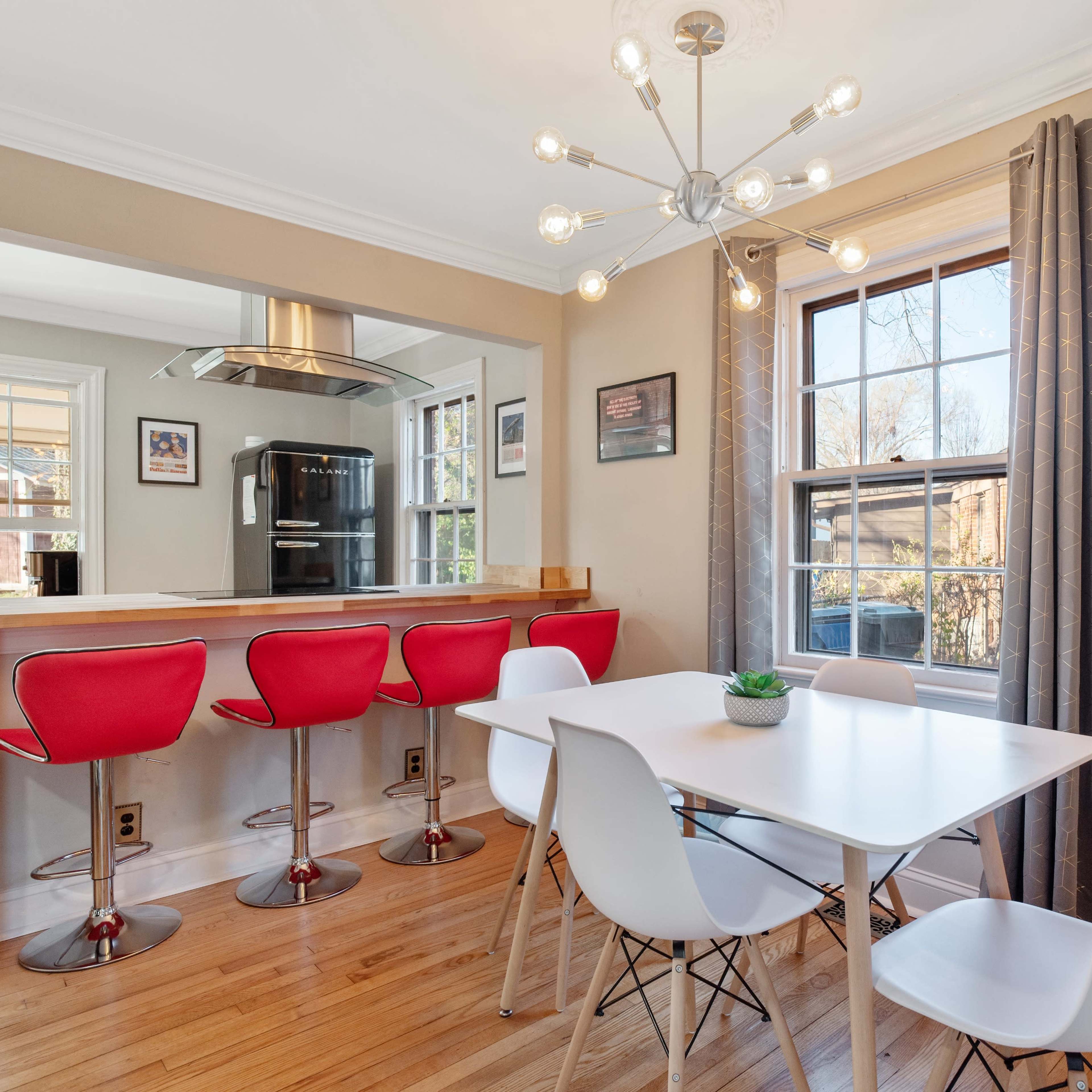 A modern dining area with a white table and chairs beside a kitchen bar with red stools, illuminated by a contemporary light fixture.