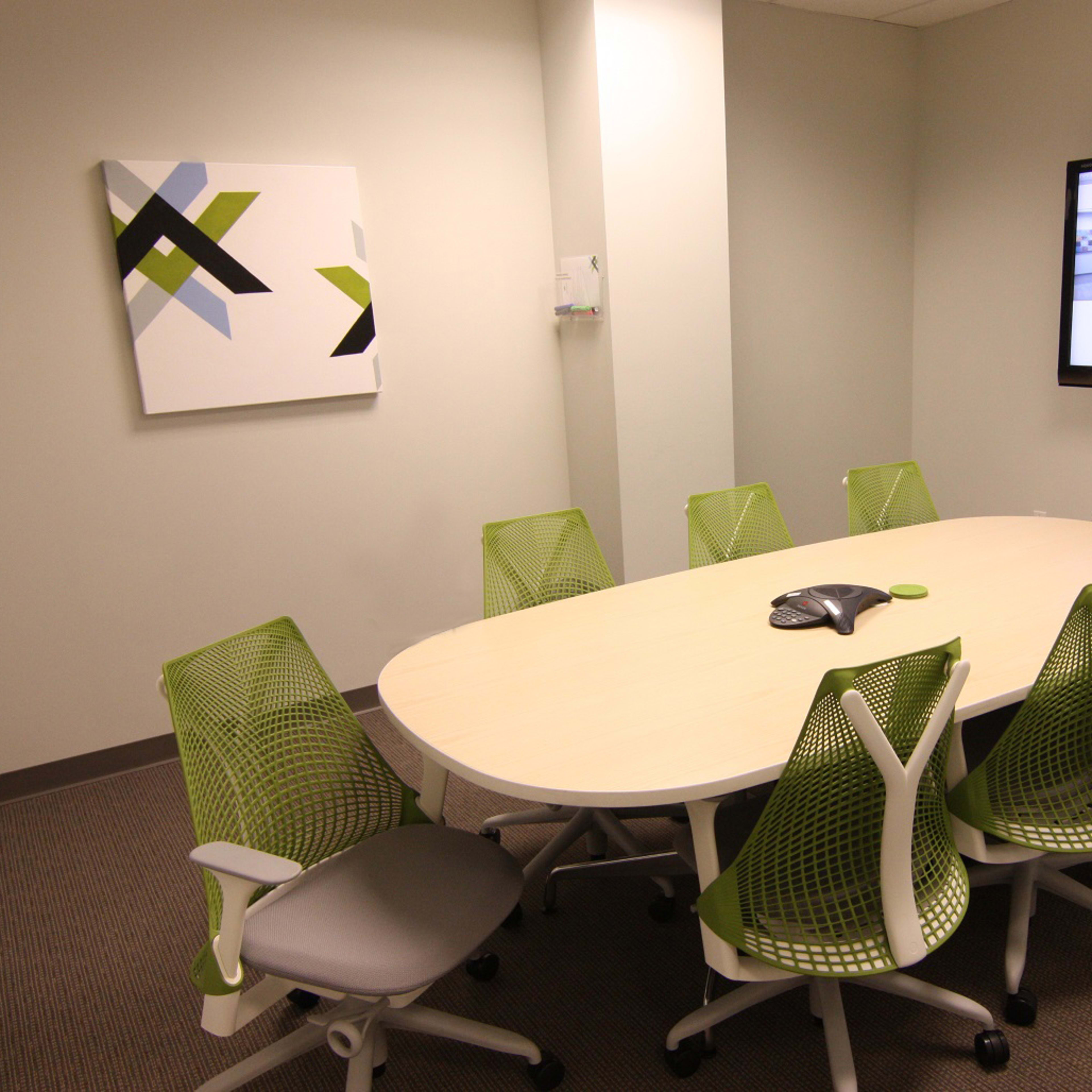 A modern conference room features a large oval table surrounded by green mesh chairs and a wall-mounted screen displaying a webpage.