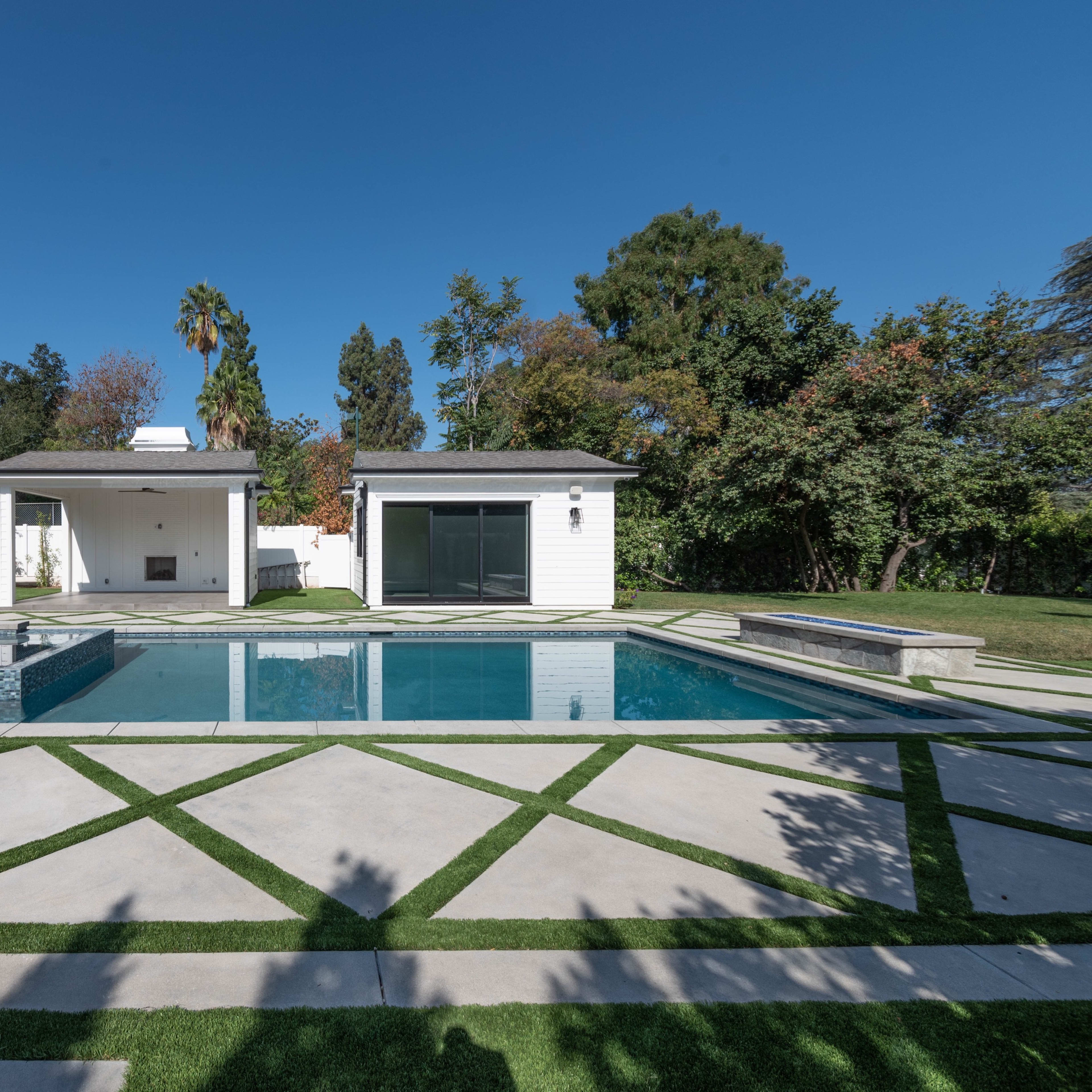 A modern backyard featuring a swimming pool surrounded by geometric stone pathways and landscaped greenery, with a shaded seating area and poolside structures.