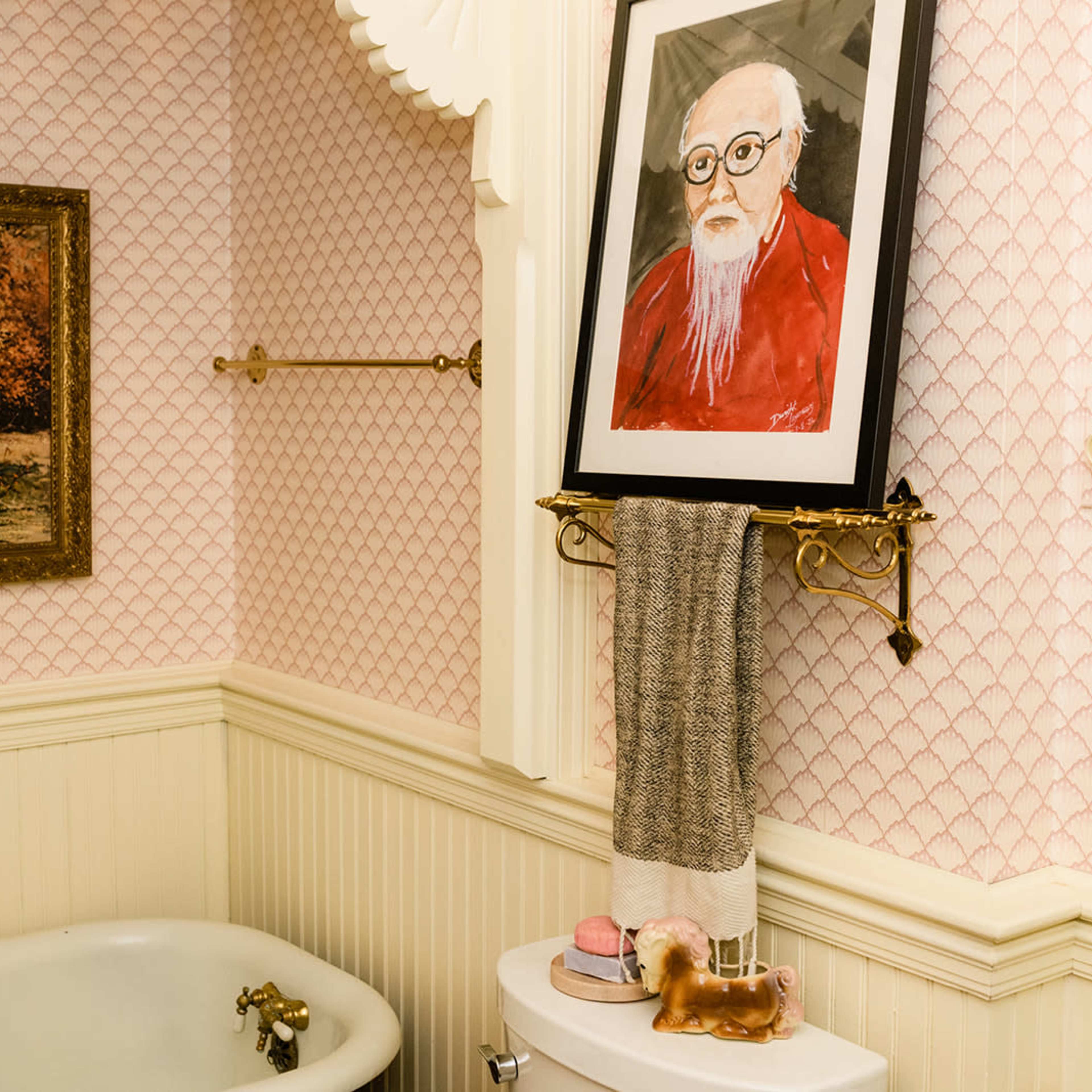 A bathroom with a vintage clawfoot tub, patterned wallpaper, and a framed portrait hung above a towel rack.