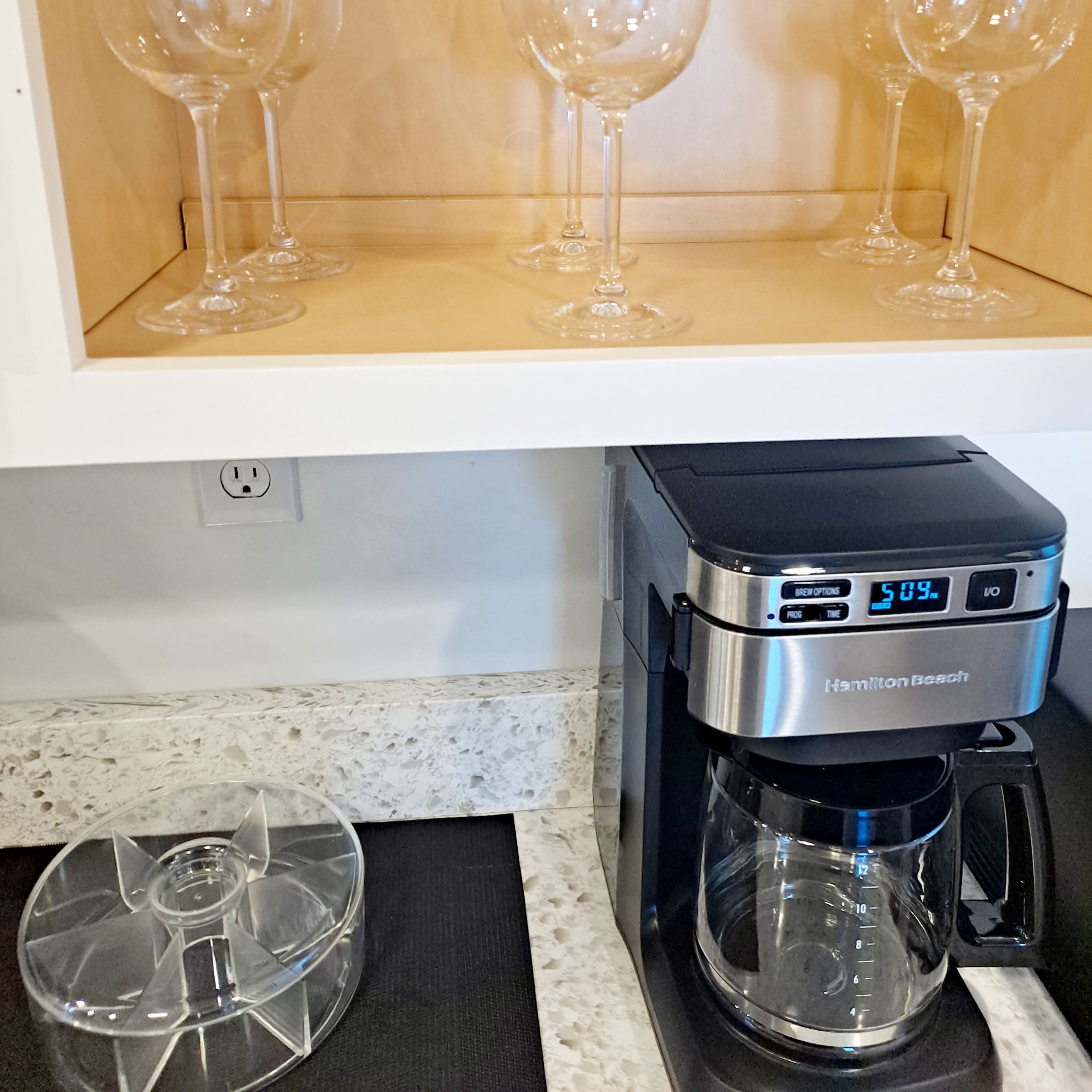 A set of clear wine glasses is neatly arranged on a wooden shelf above a Hamilton Beach coffee maker on a countertop.