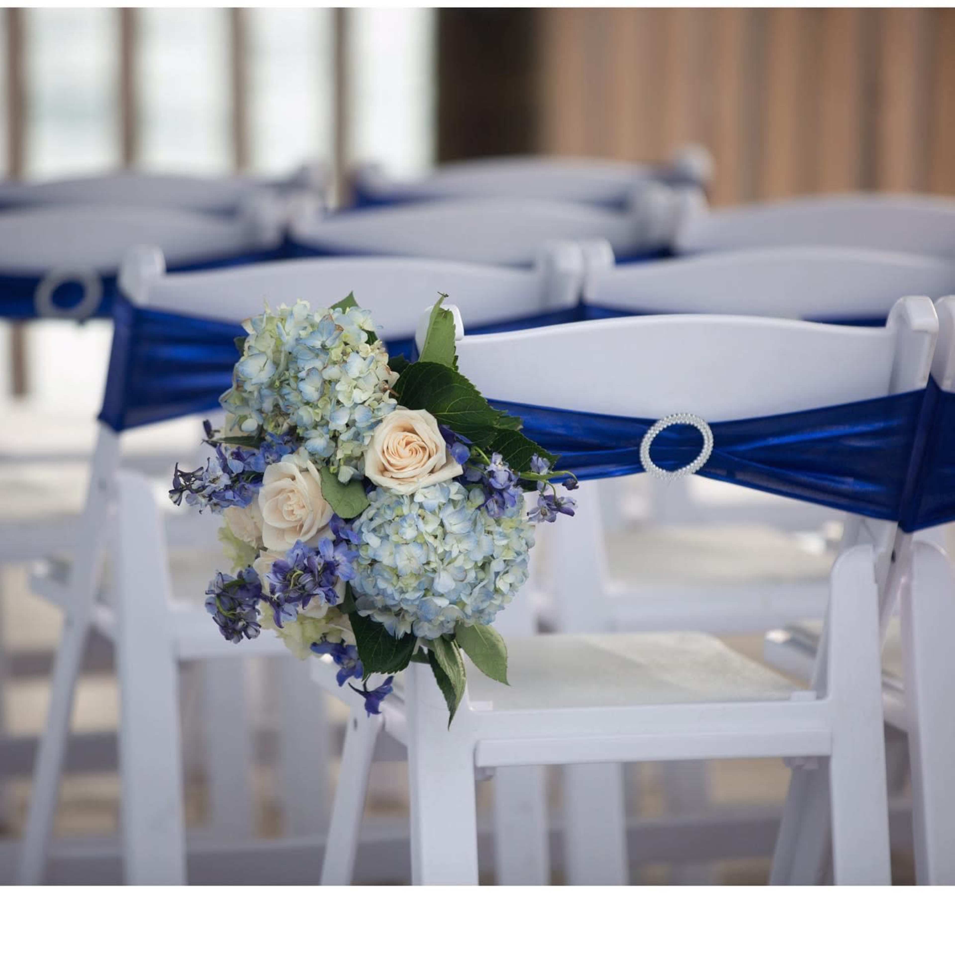 A white chair with a blue ribbon tied around the back holds a floral arrangement of blue and cream flowers.