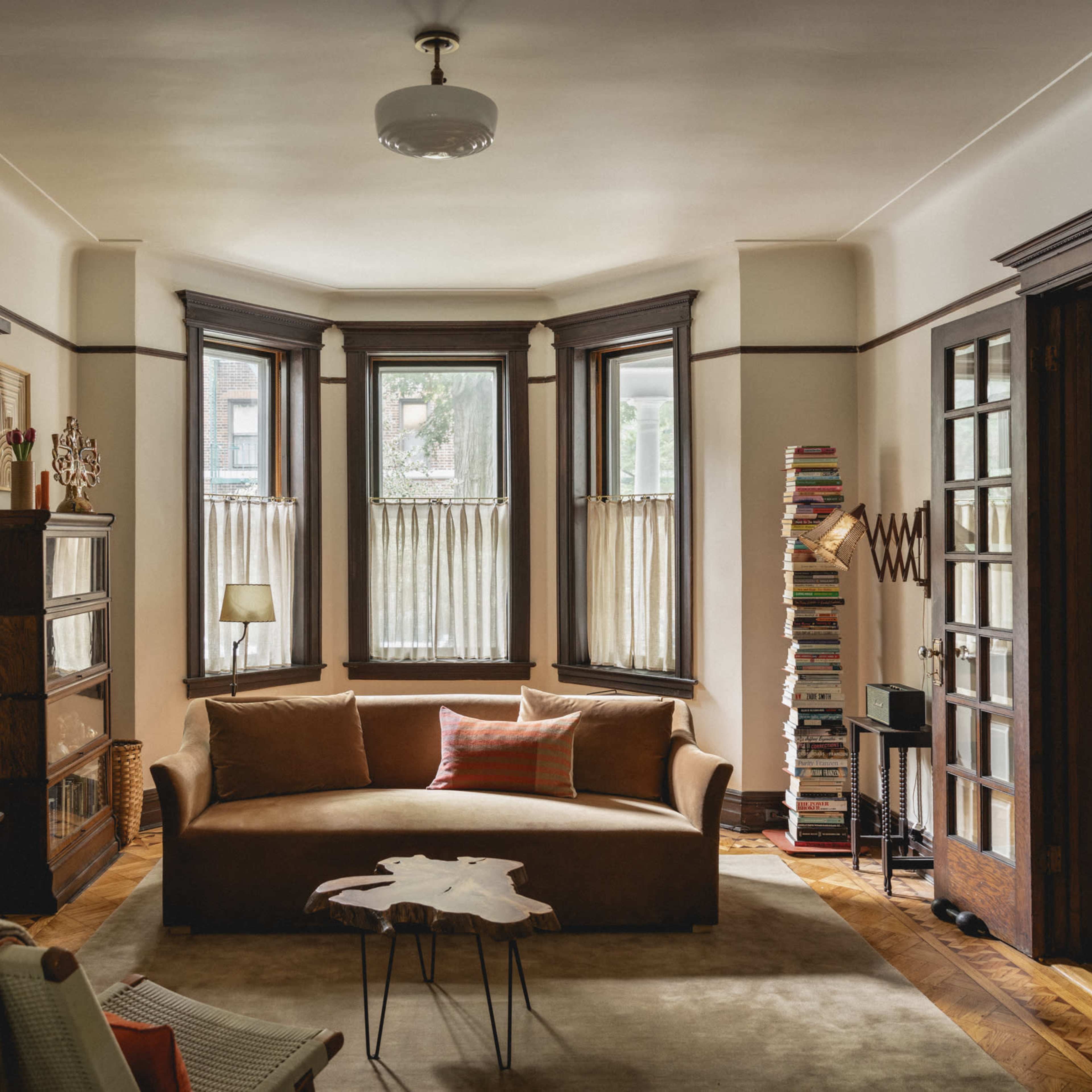 A living room with a brown sofa, an area rug, large windows with sheer curtains, and bookshelves filled with books.