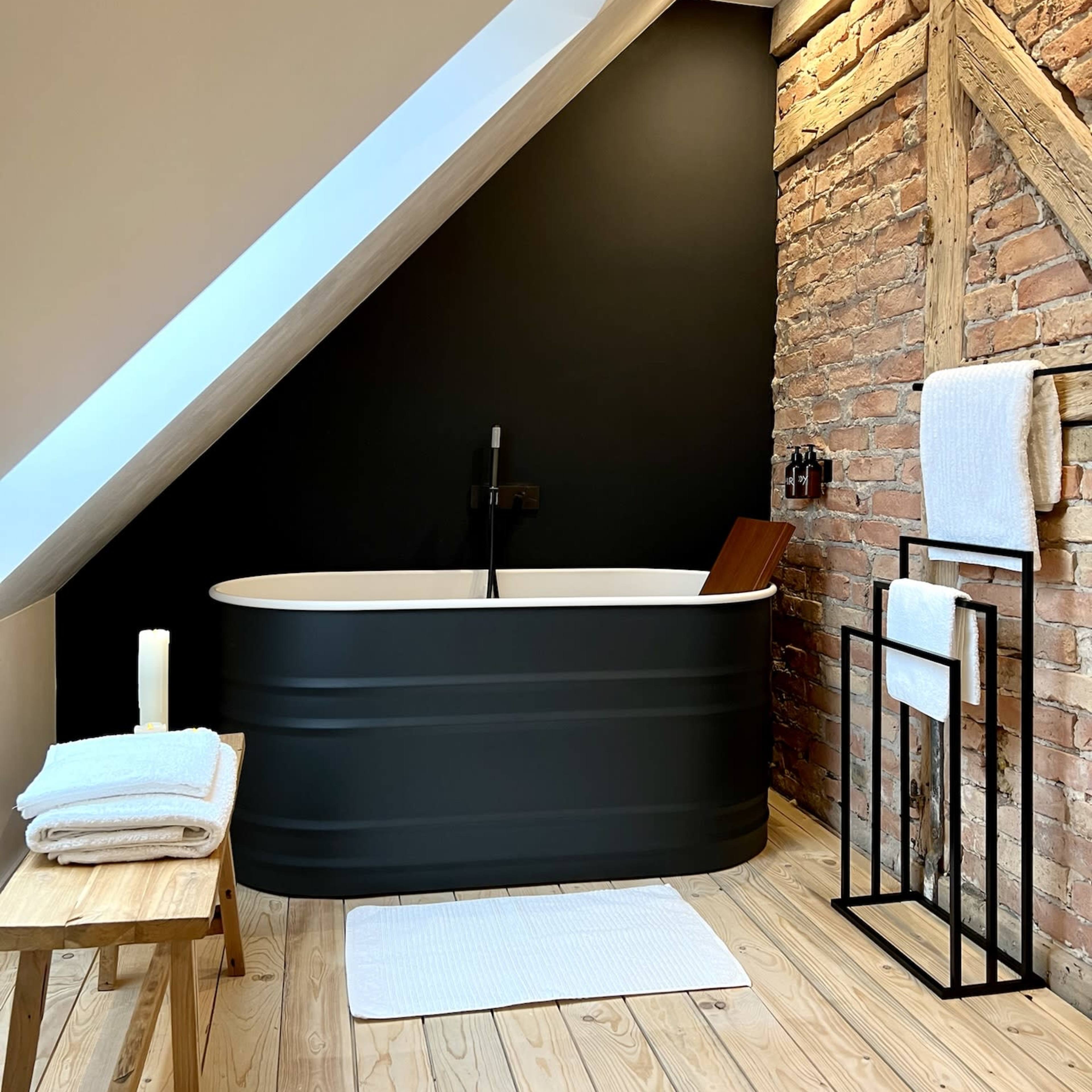 A modern bathroom featuring a black freestanding tub against a brick wall, wooden flooring, and neatly arranged towels.