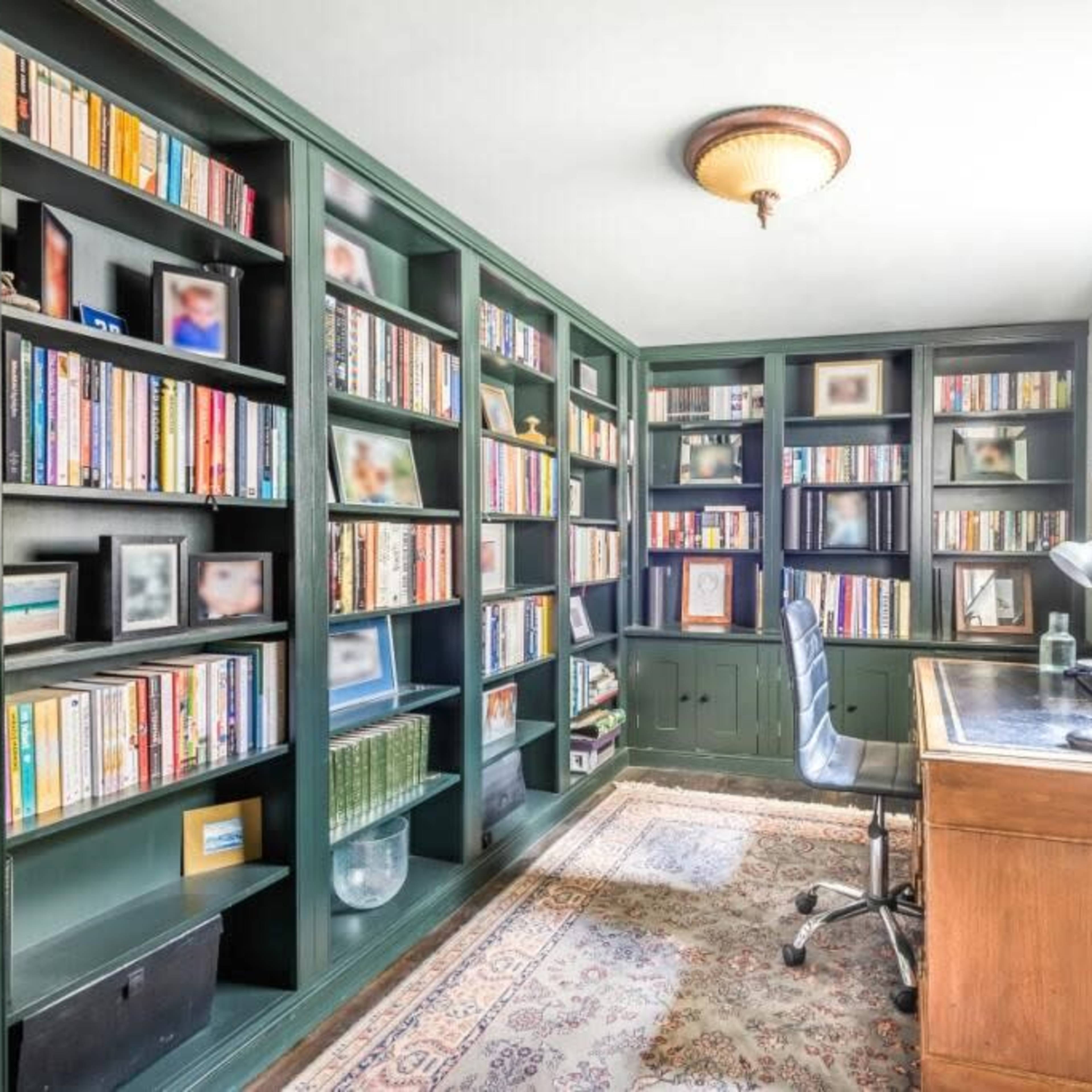 A small room features green bookshelves filled with books, a wooden desk with a computer, and a patterned rug on the floor.