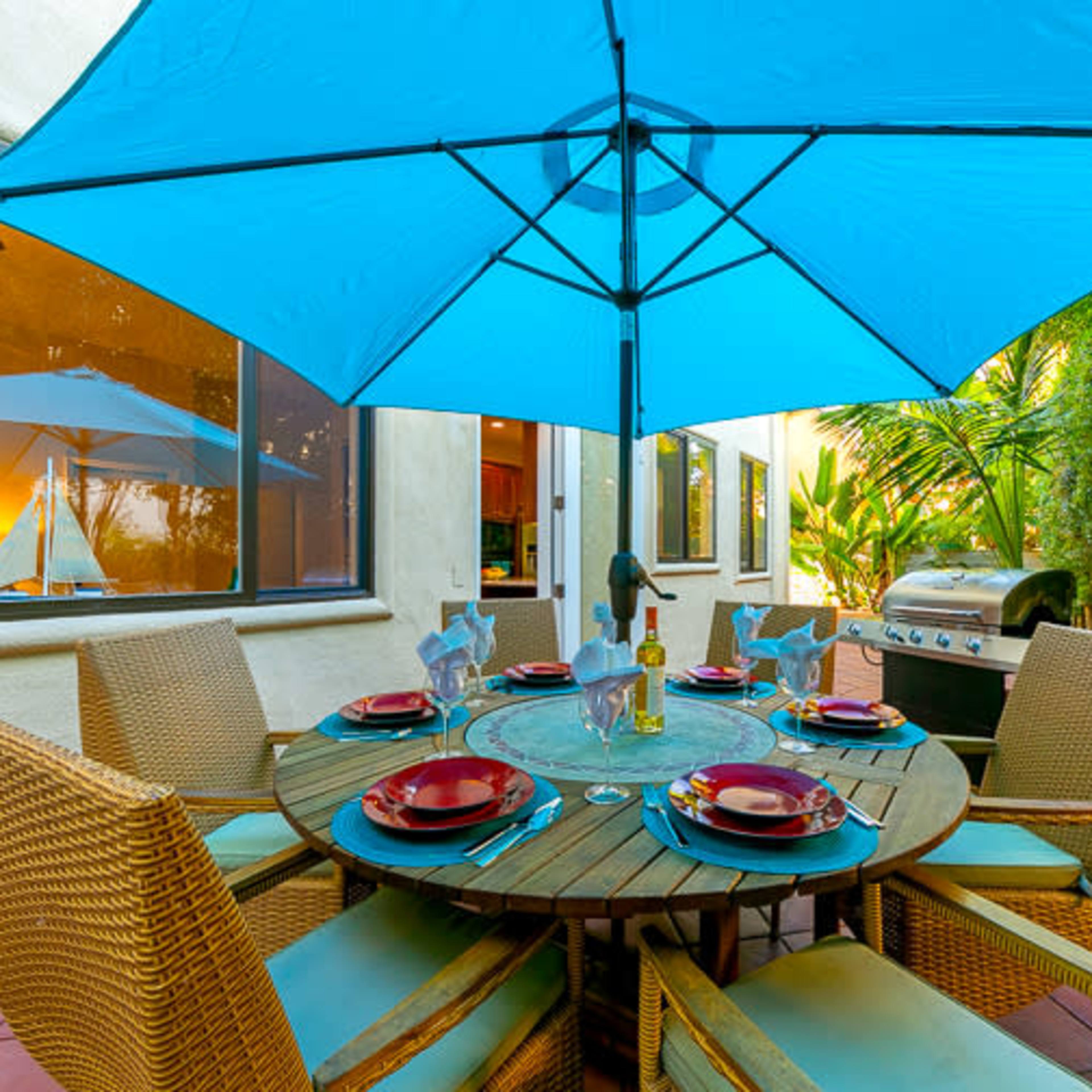 A shaded outdoor dining table set with plates and chairs is positioned near a barbecue grill and surrounded by lush greenery.