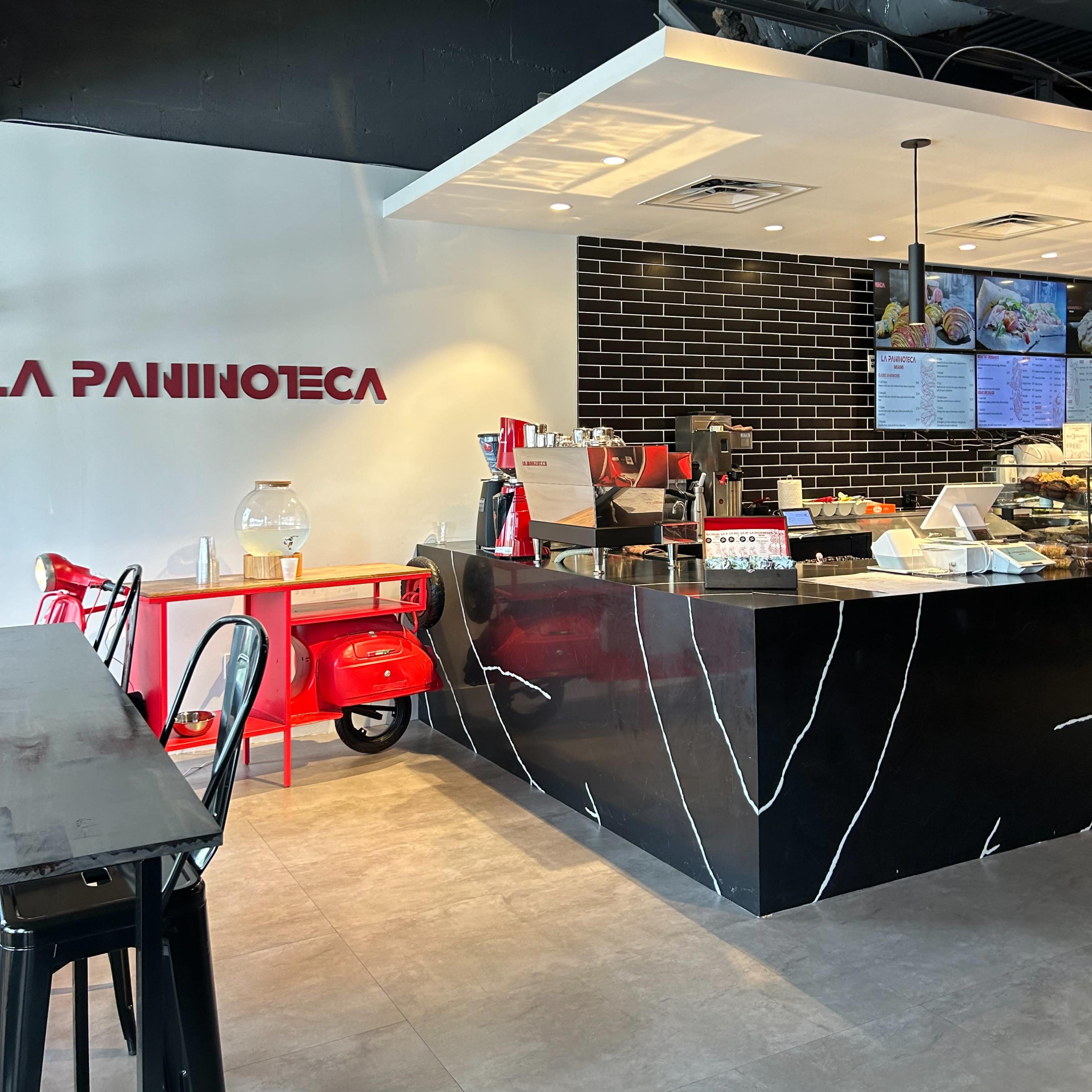 The interior of a café with a counter featuring a display of food, a coffee machine, and a red scooter parked nearby.