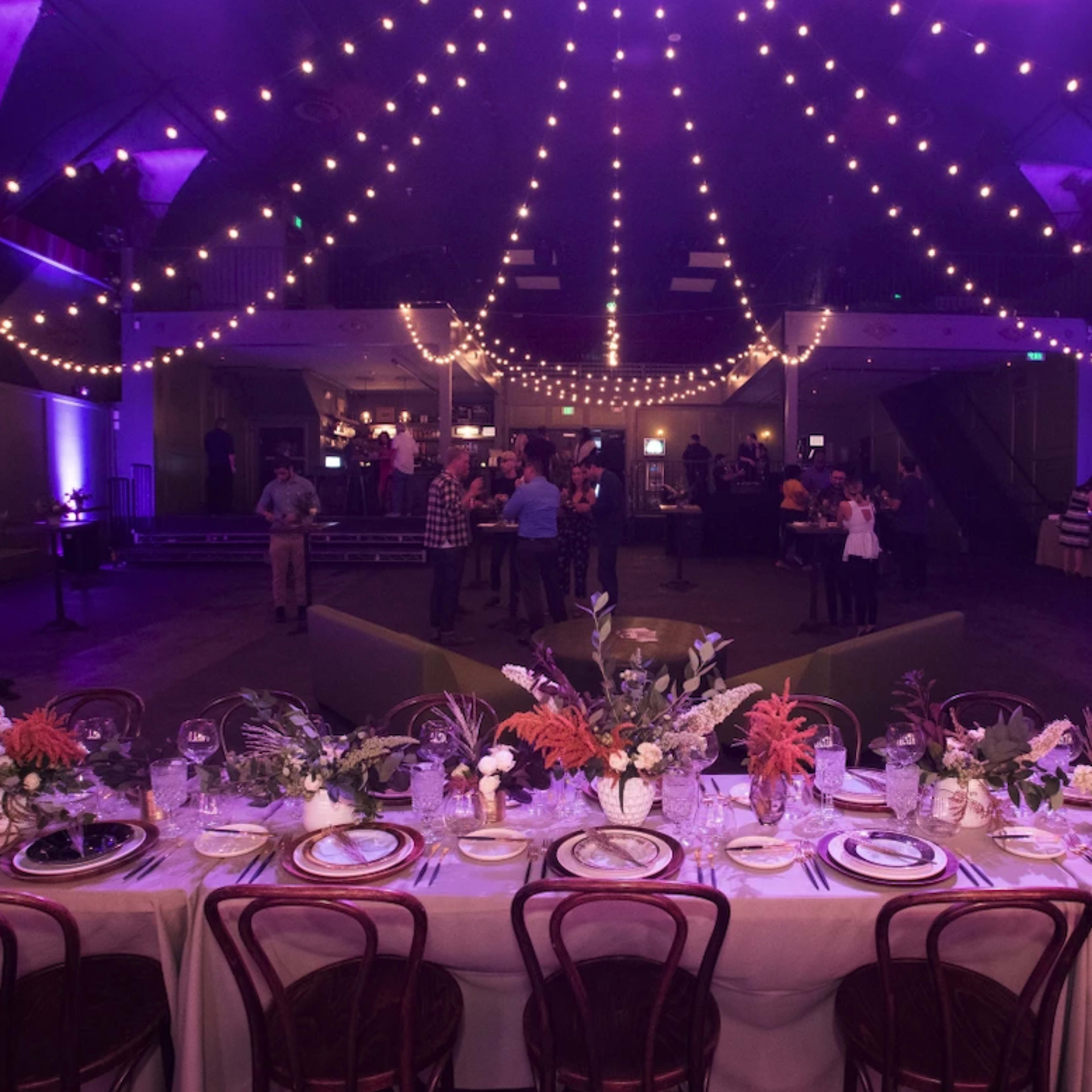 A long dining table is set with floral centerpieces and elegant dinnerware under a canopy of string lights in a warmly lit event space.