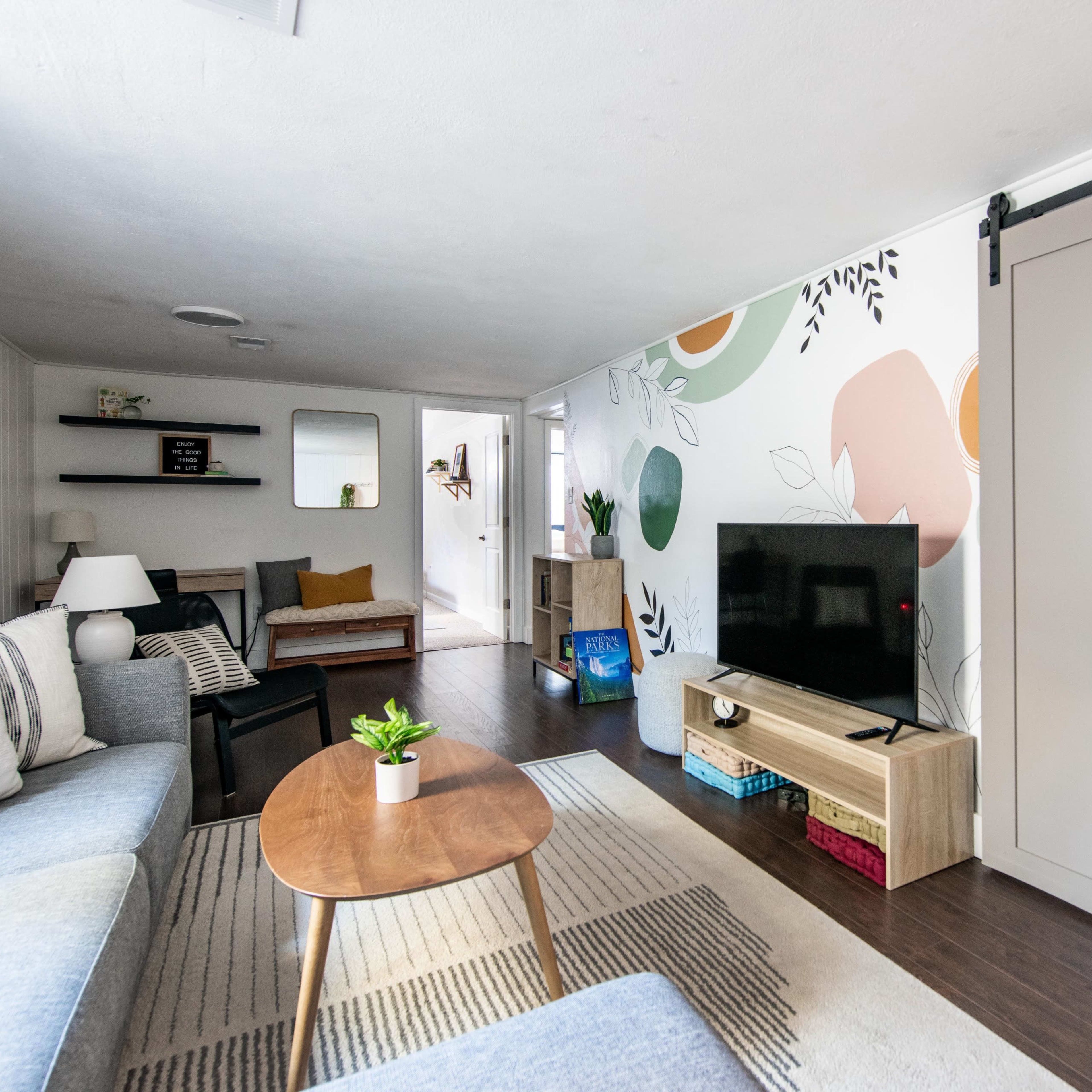 A living room features a couch, a wooden table, a television on a stand, and a wall with colorful plant-themed murals.