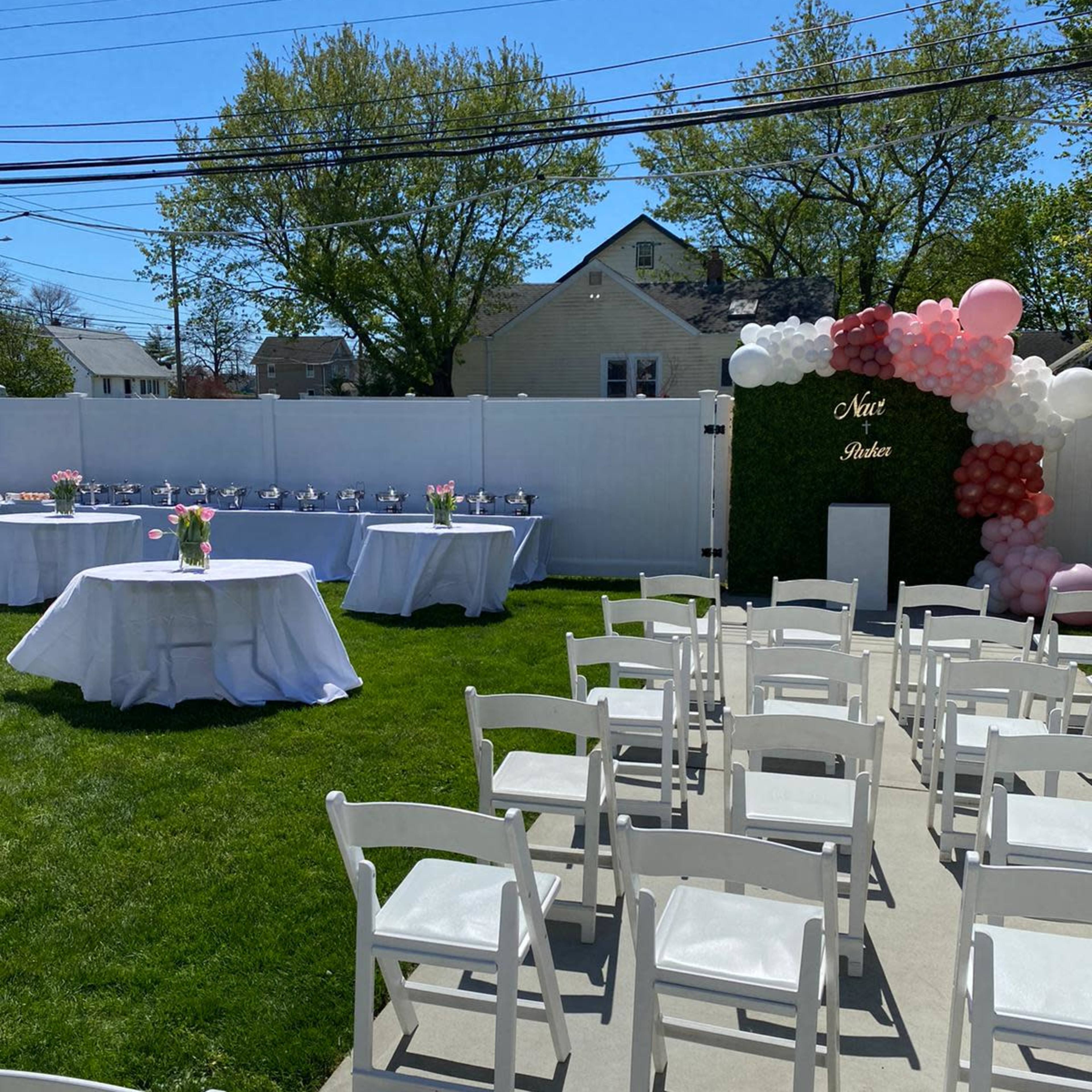 A lawn is set up with white chairs arranged facing a table decorated with flowers, alongside a wall adorned with balloons for a celebration.