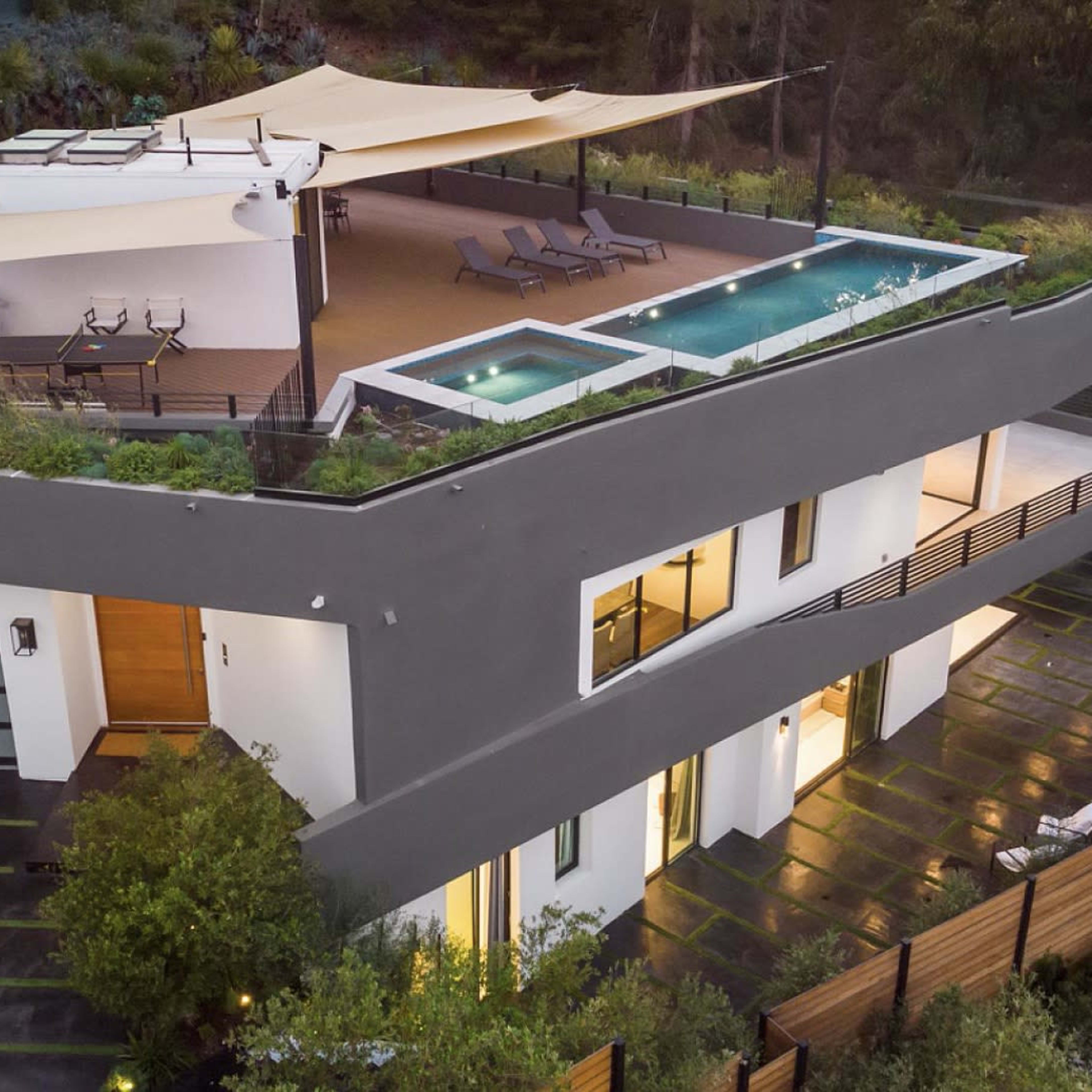 A modern two-story house features a rooftop deck with a pool and seating area, surrounded by greenery and a paved driveway.
