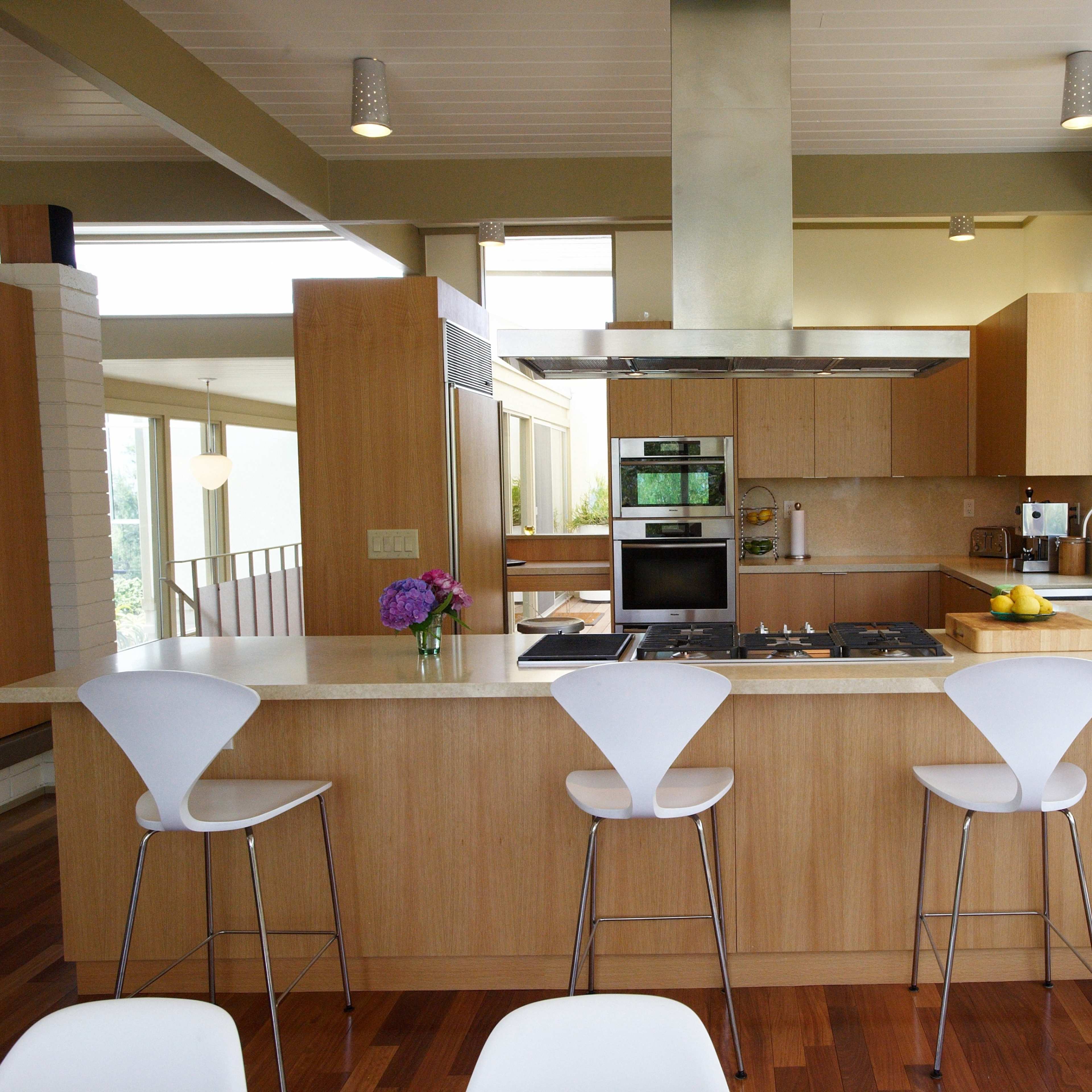 A modern kitchen features a central island with three white stools, stainless steel appliances, and large windows overlooking greenery.