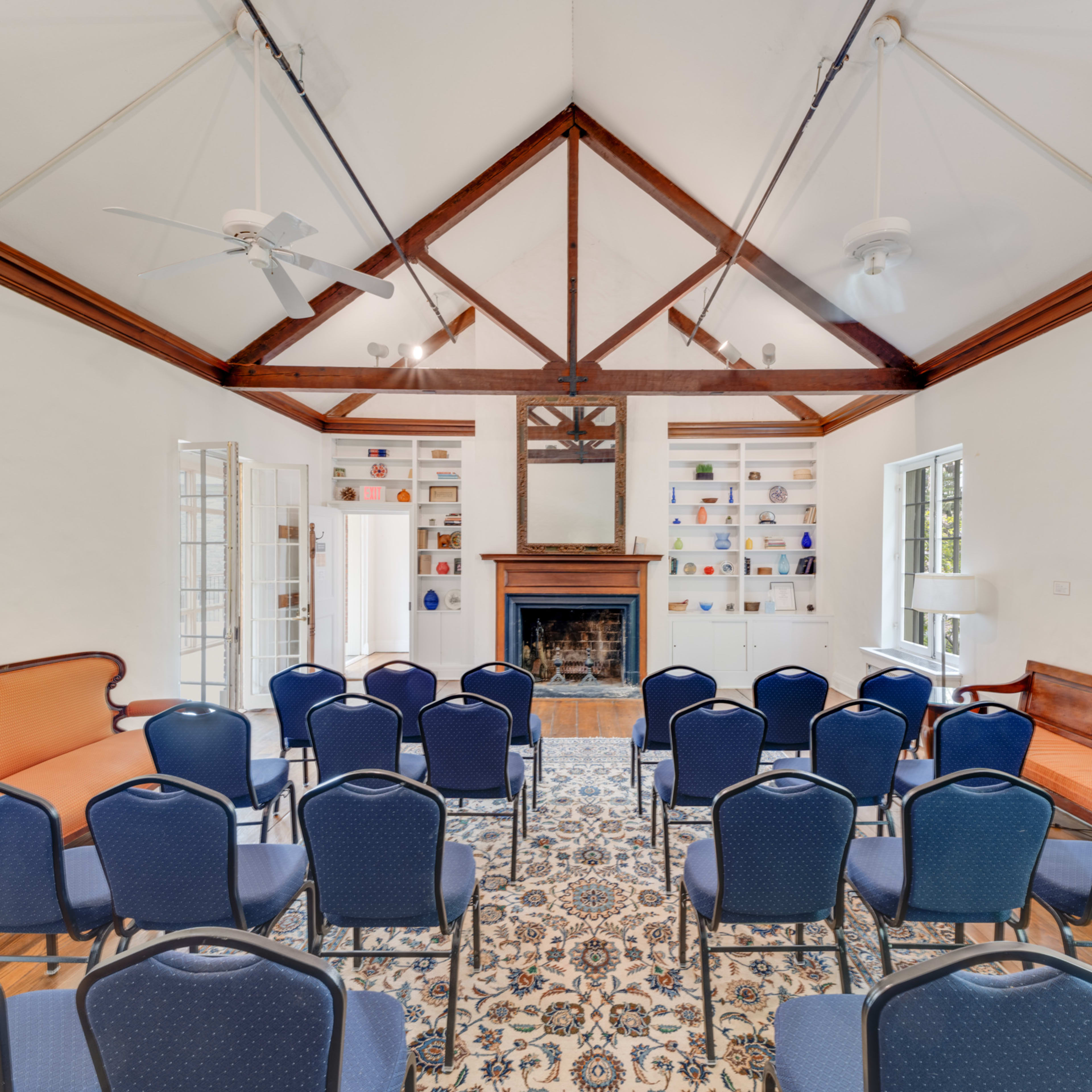 A seminar room arranged with several blue chairs facing a central fireplace, framed by a high ceiling with exposed wooden beams and shelves displaying items on the wall.
