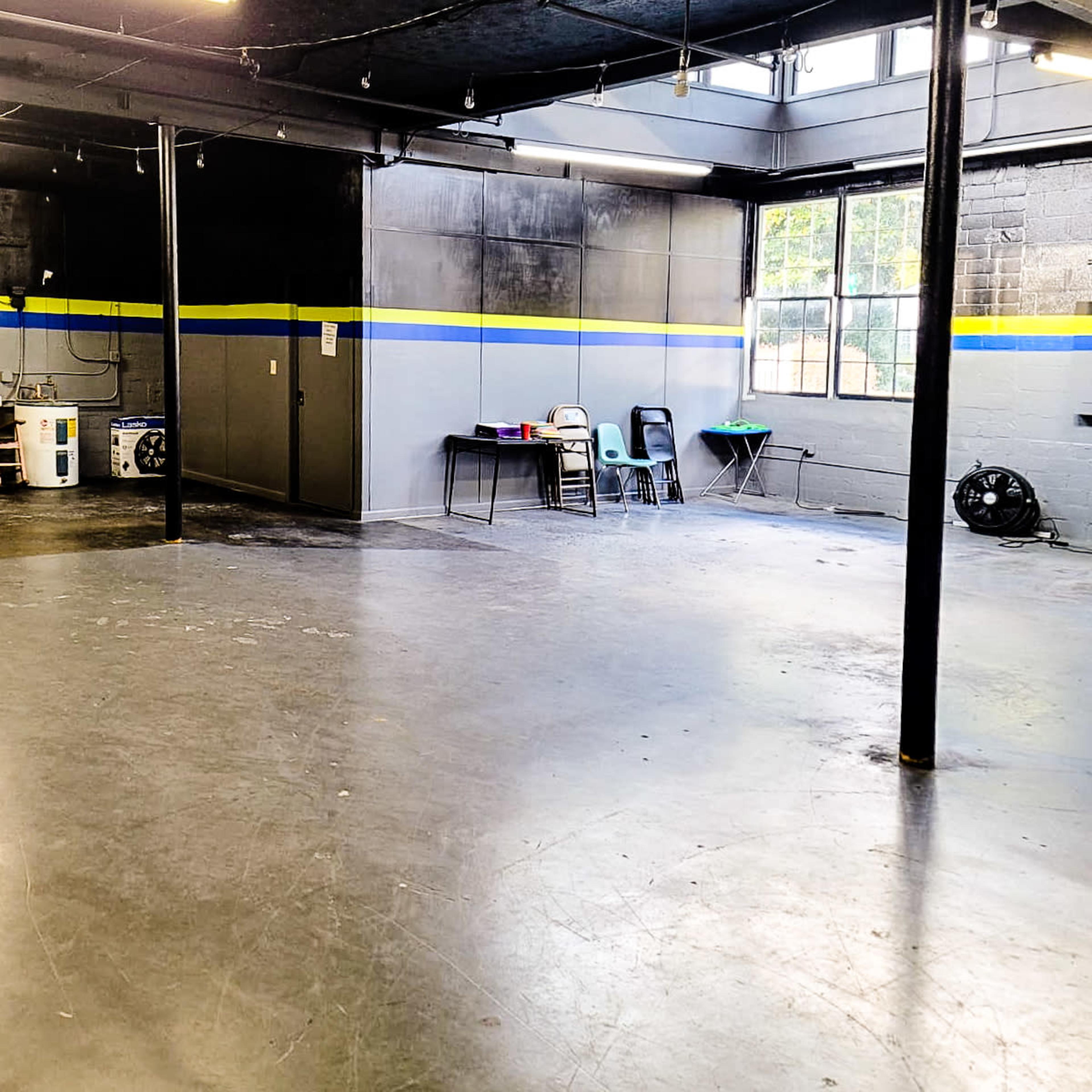A spacious, vacant interior room with a polished concrete floor, a table with chairs in one corner, and large windows letting in natural light.