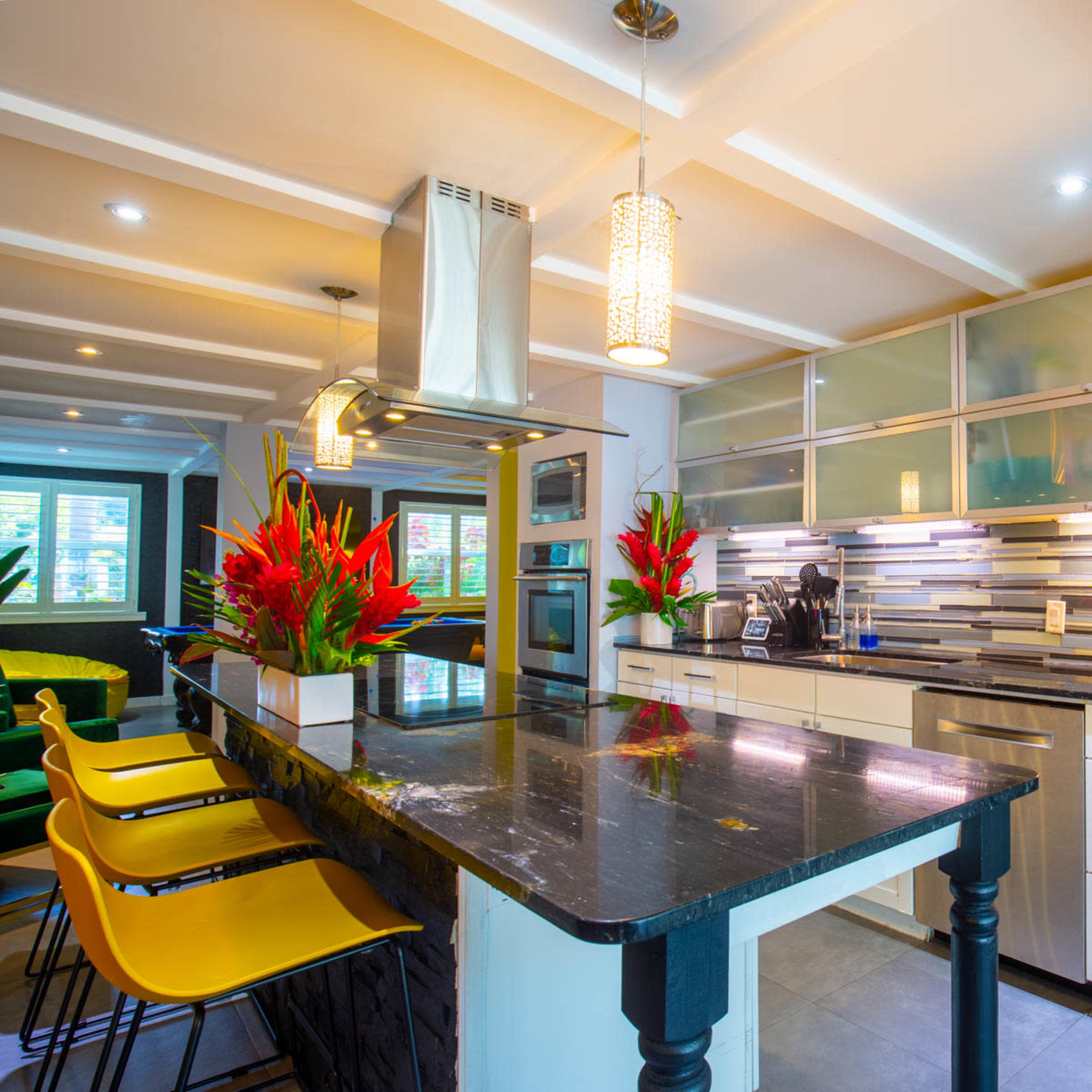 A modern kitchen features a central island with yellow bar stools, stainless steel appliances, and vibrant plants against a backdrop of sleek cabinetry and decorative lighting.