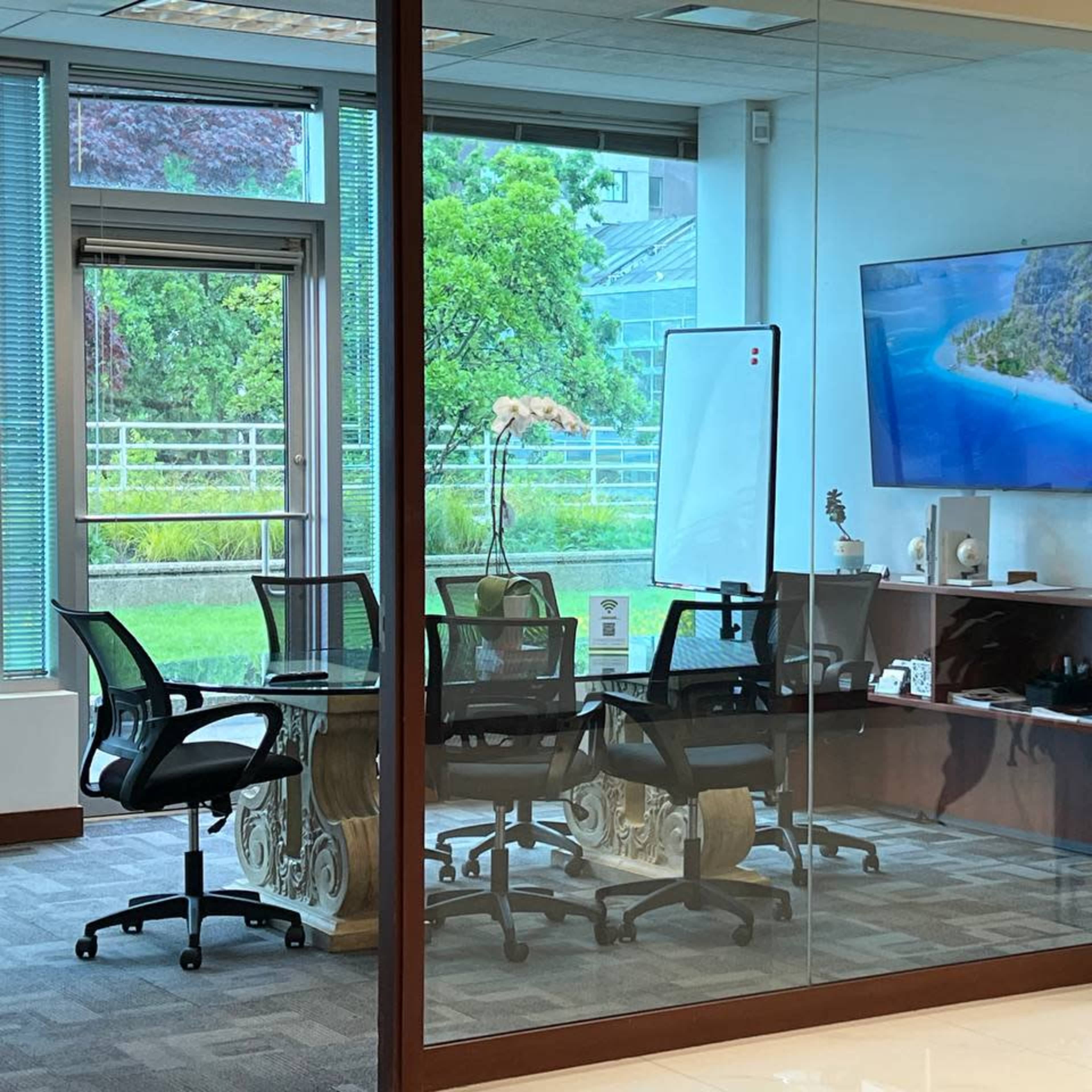 A modern conference room features glass walls, a large screen displaying a scenic image, and a round table surrounded by black chairs.