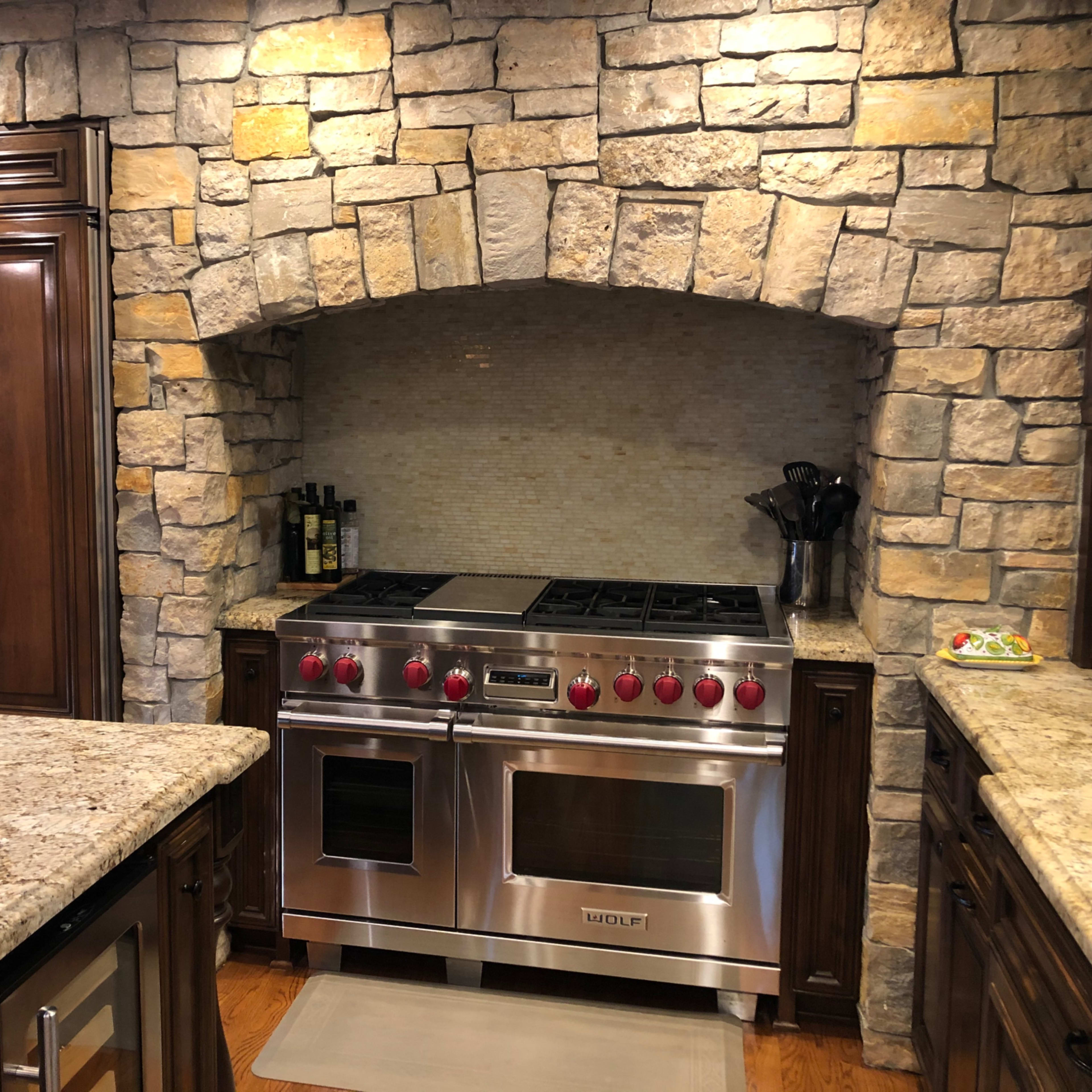 A kitchen featuring a stainless steel range with red knobs, set against a rustic stone arch and dark wood cabinetry.