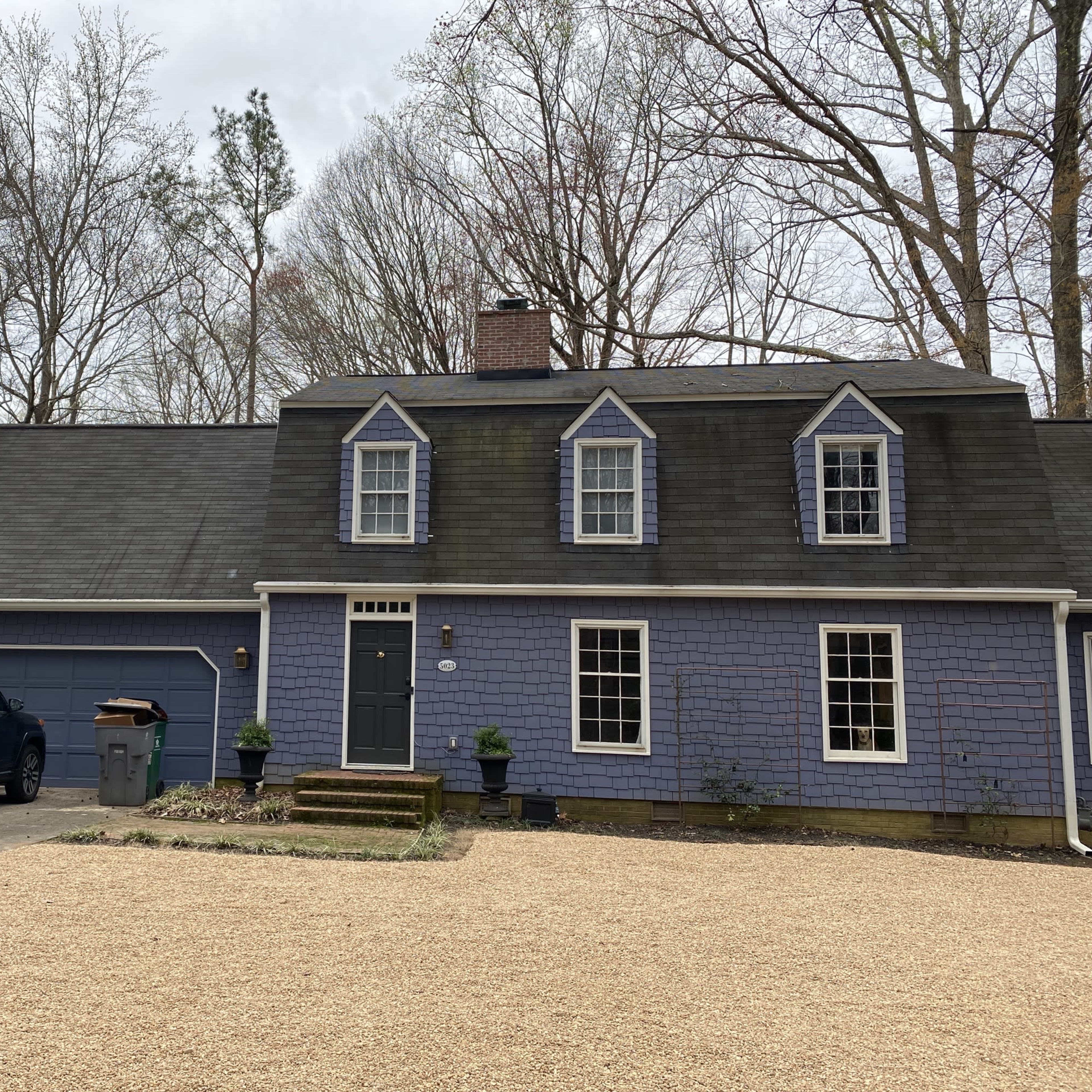 A two-story, blue house with a front porch and a gravel driveway is surrounded by trees.