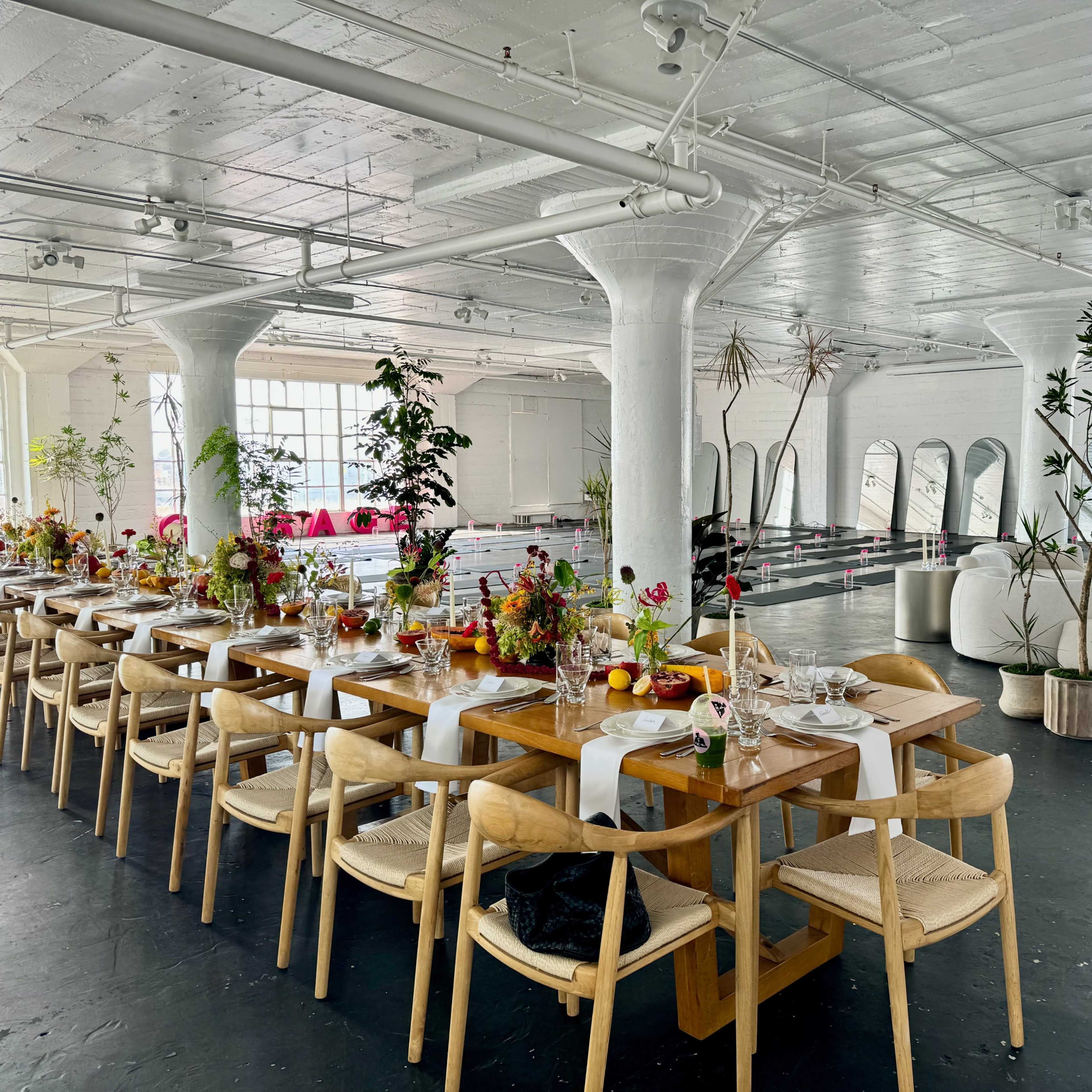 A long wooden dining table is set up in a spacious, brightly lit room with large windows, decorated with floral centerpieces and surrounded by modern seating.