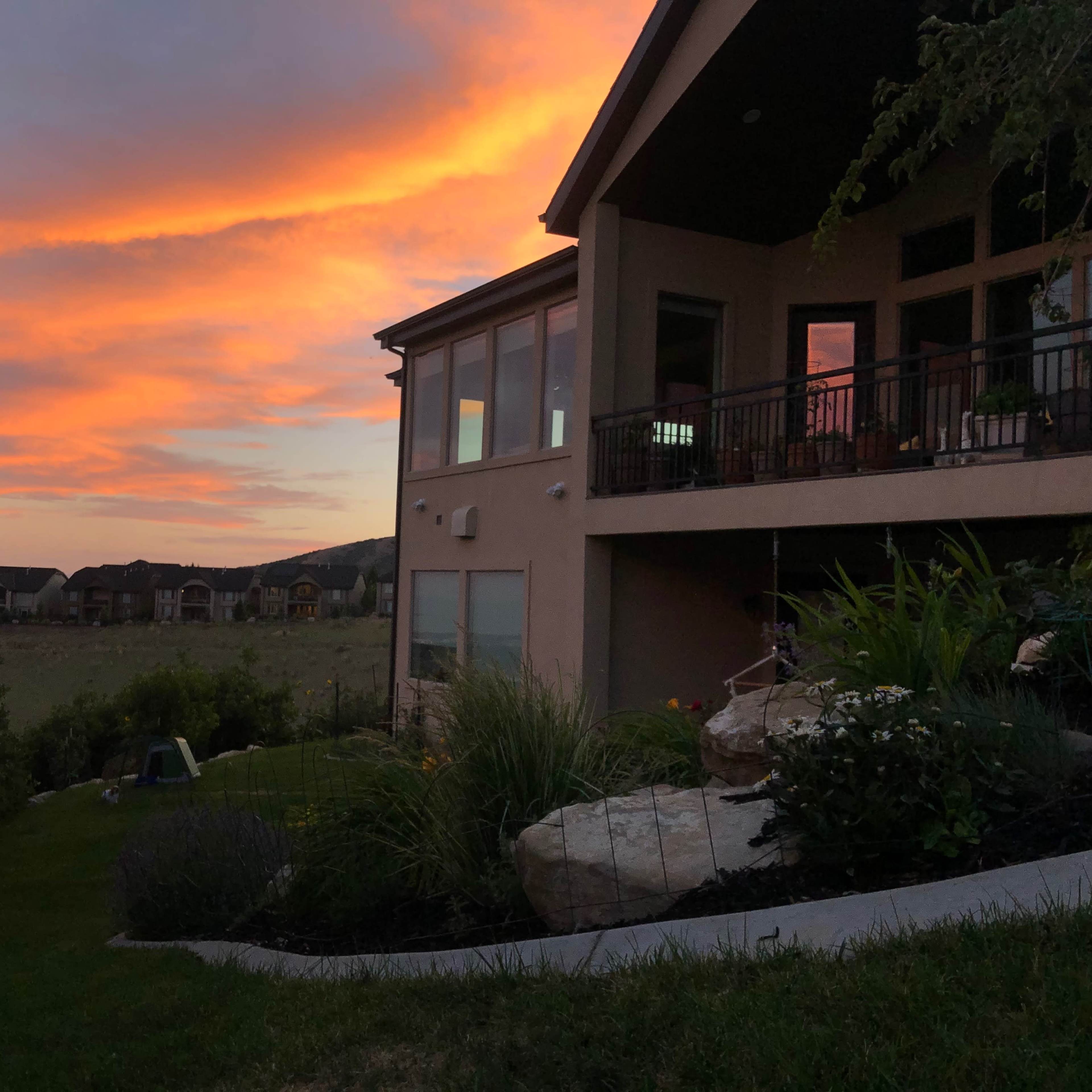 A two-story house with a balcony overlooks a grassy area and a sunset sky filled with orange and pink hues.
