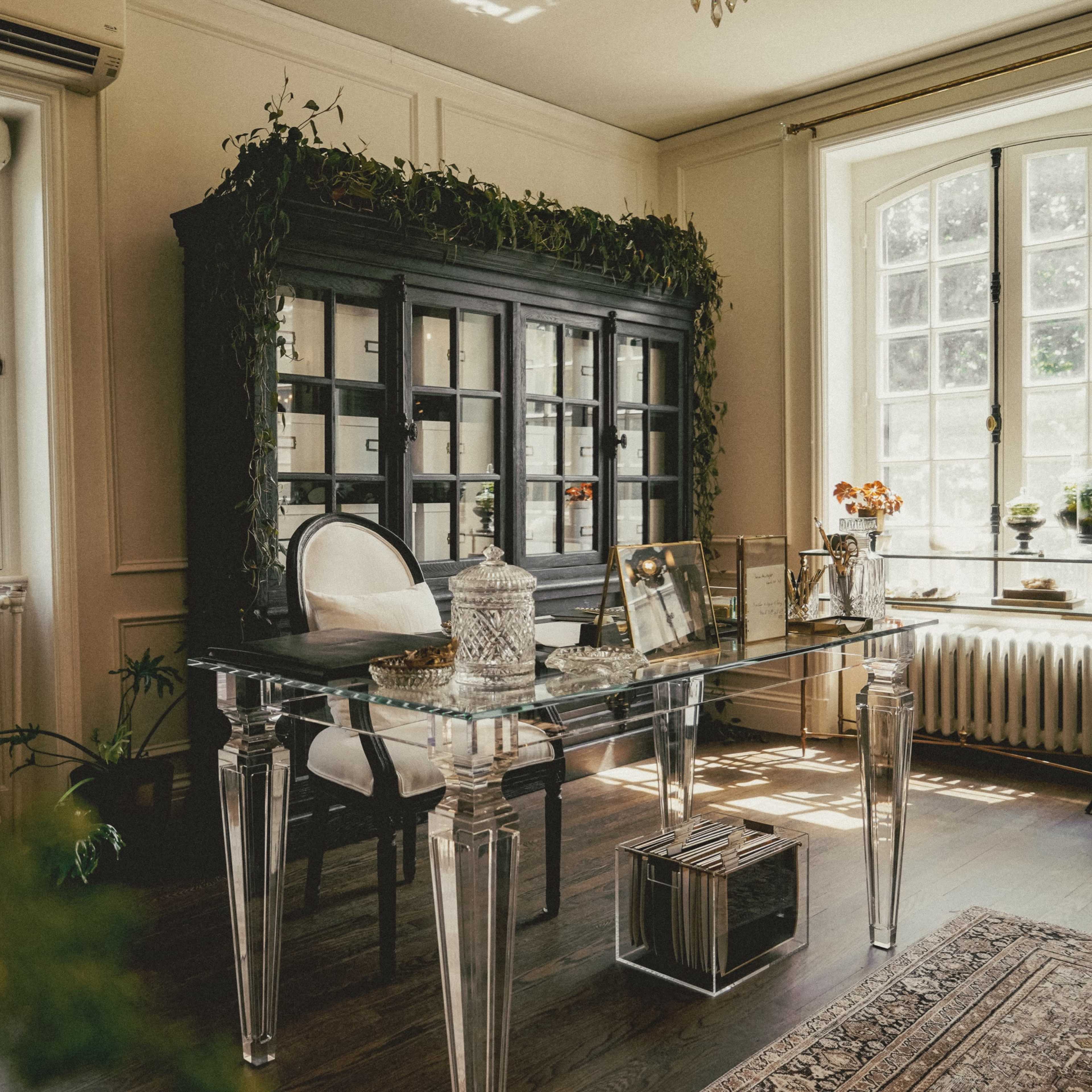 A home office features a glass desk, an ornate black cabinet surrounded by plants, and large windows providing natural light.