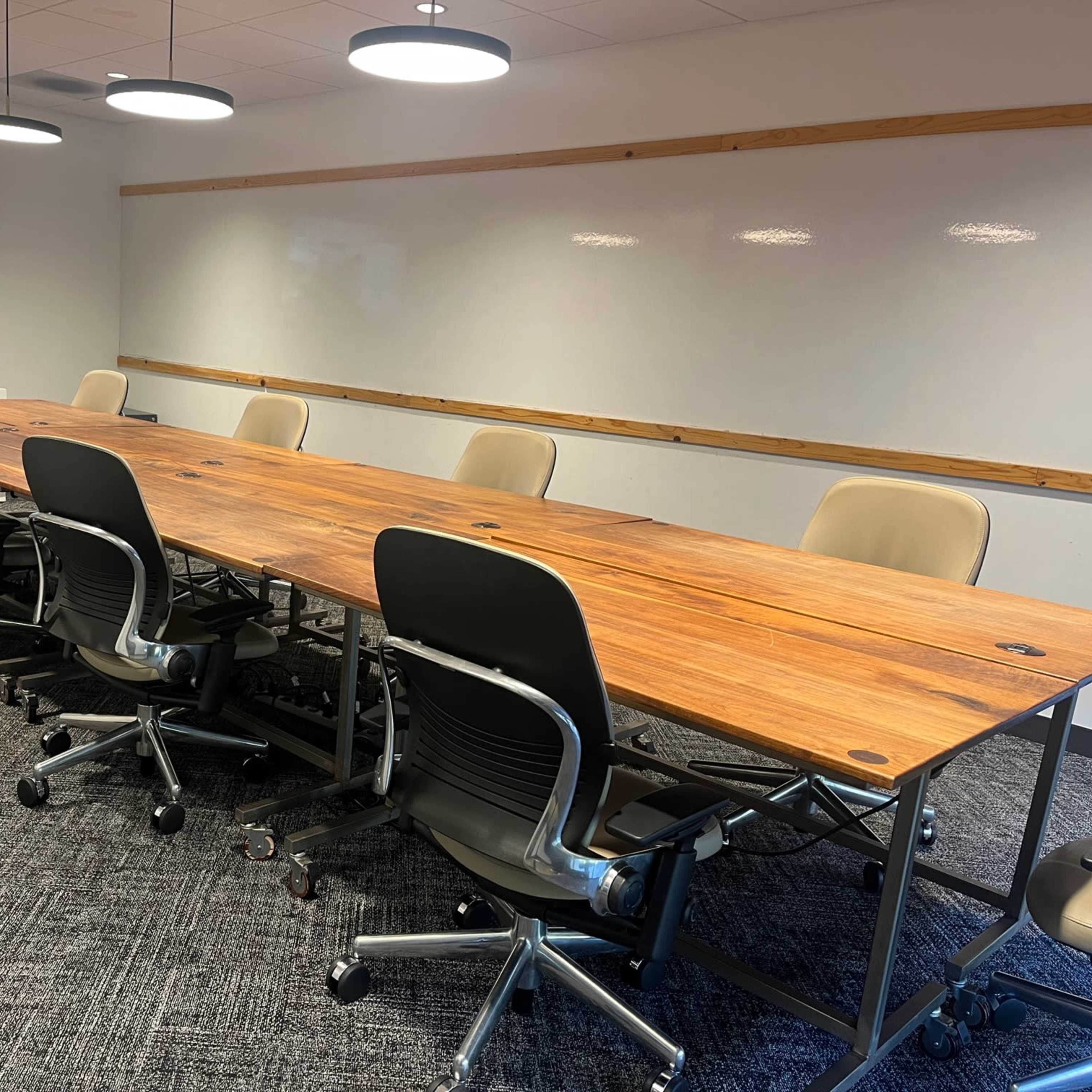 A modern conference room features a long wooden table surrounded by ergonomic chairs, with a large screen mounted on the wall and potted plants in the background.