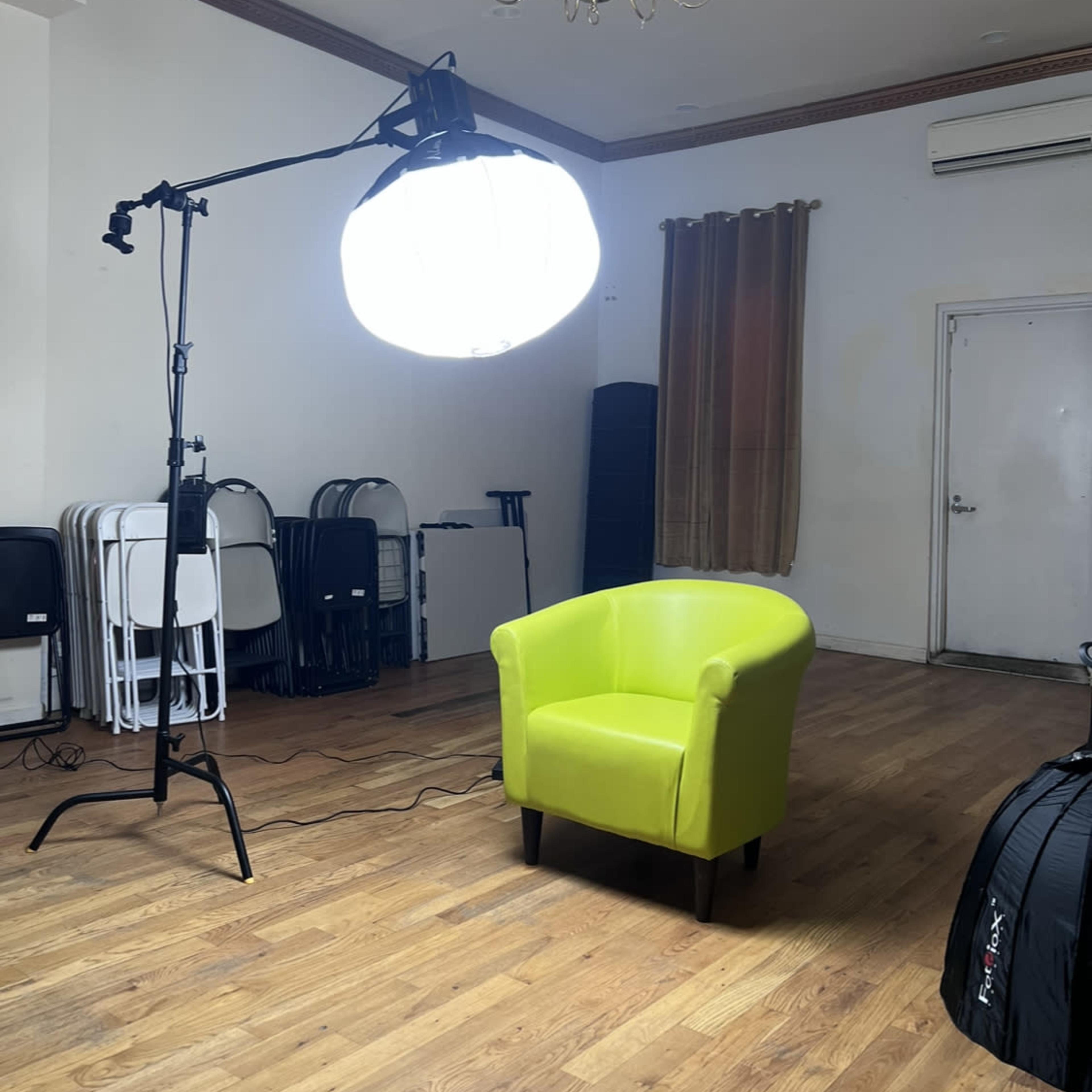 A bright green chair placed in an empty room illuminated by a large softbox light, with folding chairs stacked in the background.