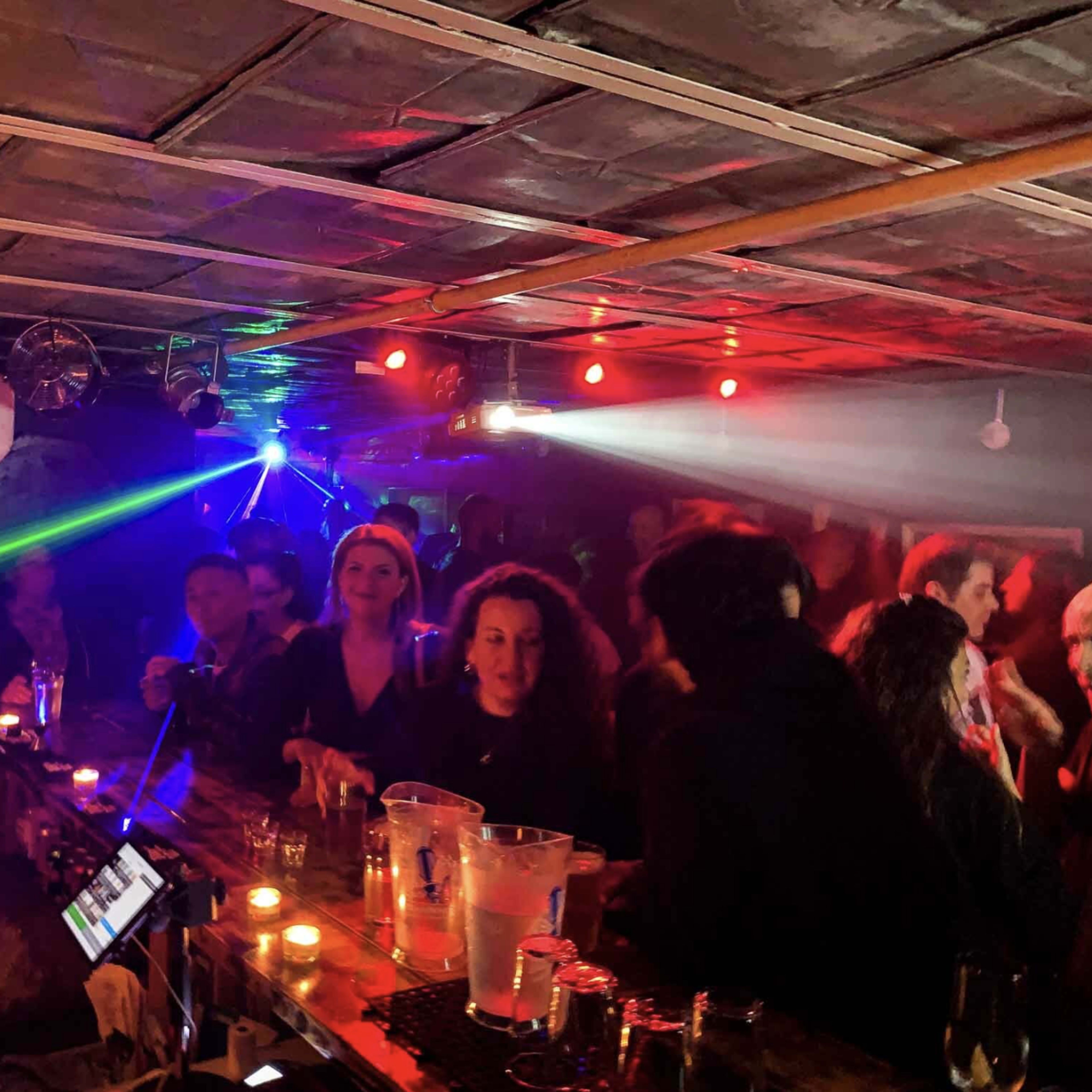 A crowded bar scene features patrons socializing under colorful lights while a bartender prepares drinks.