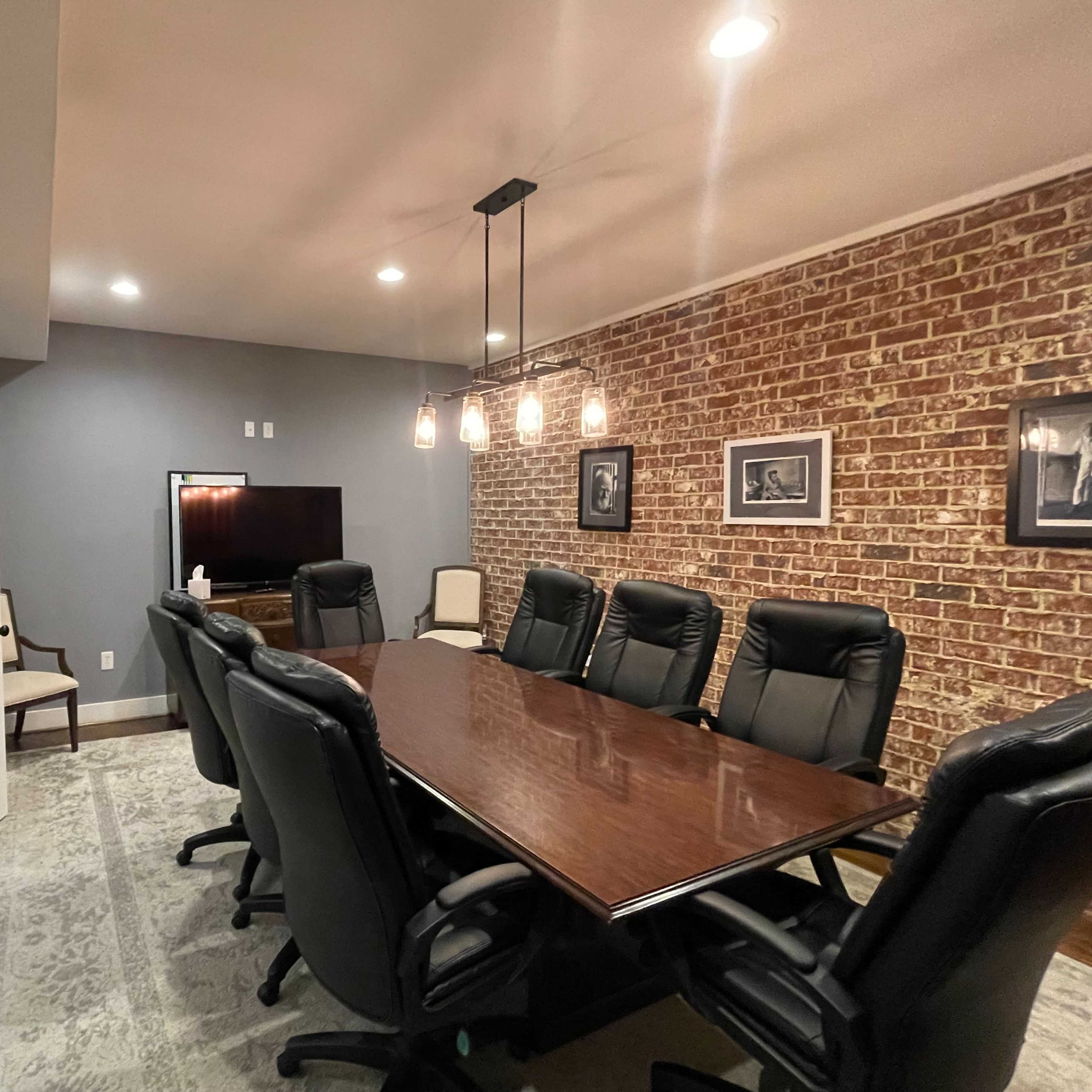 A conference room with a long wooden table surrounded by black leather chairs, a television mounted on the wall, and exposed brick accents.