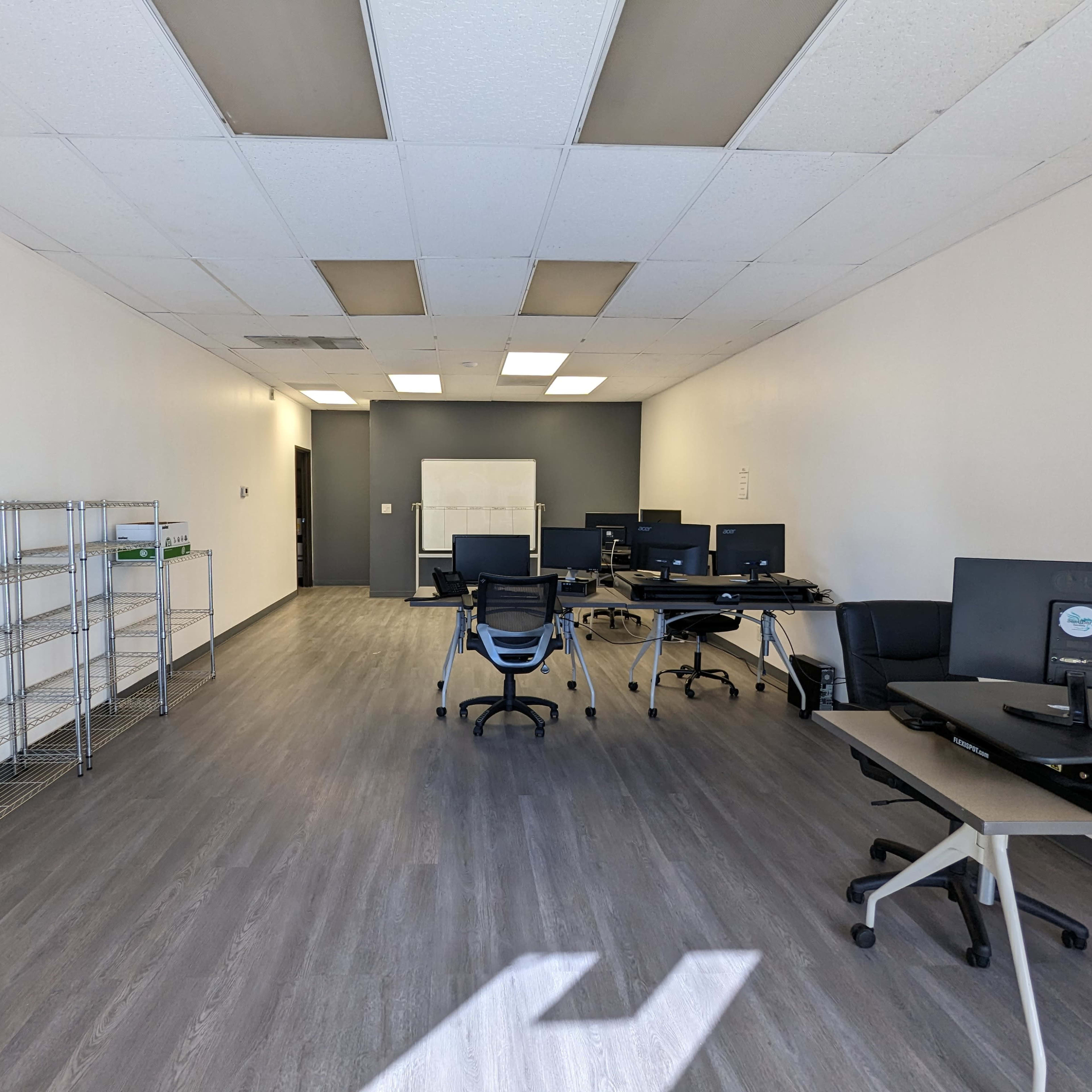 An empty office space with several computers on desks, shelves along one wall, and a whiteboard in the background.
