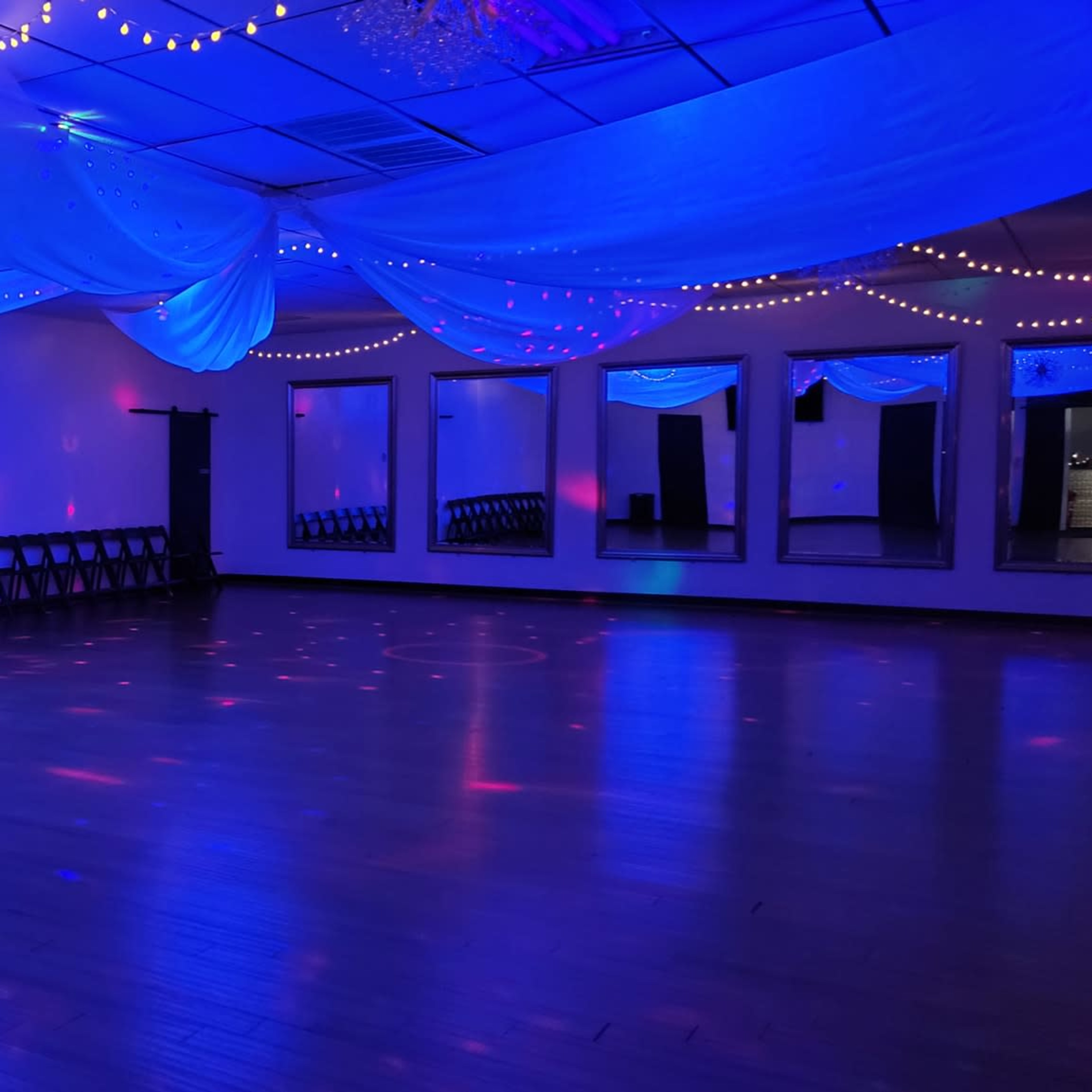 A spacious dance studio with wooden floors, decorated with blue lighting and draped fabric overhead, alongside a row of chairs against one wall.