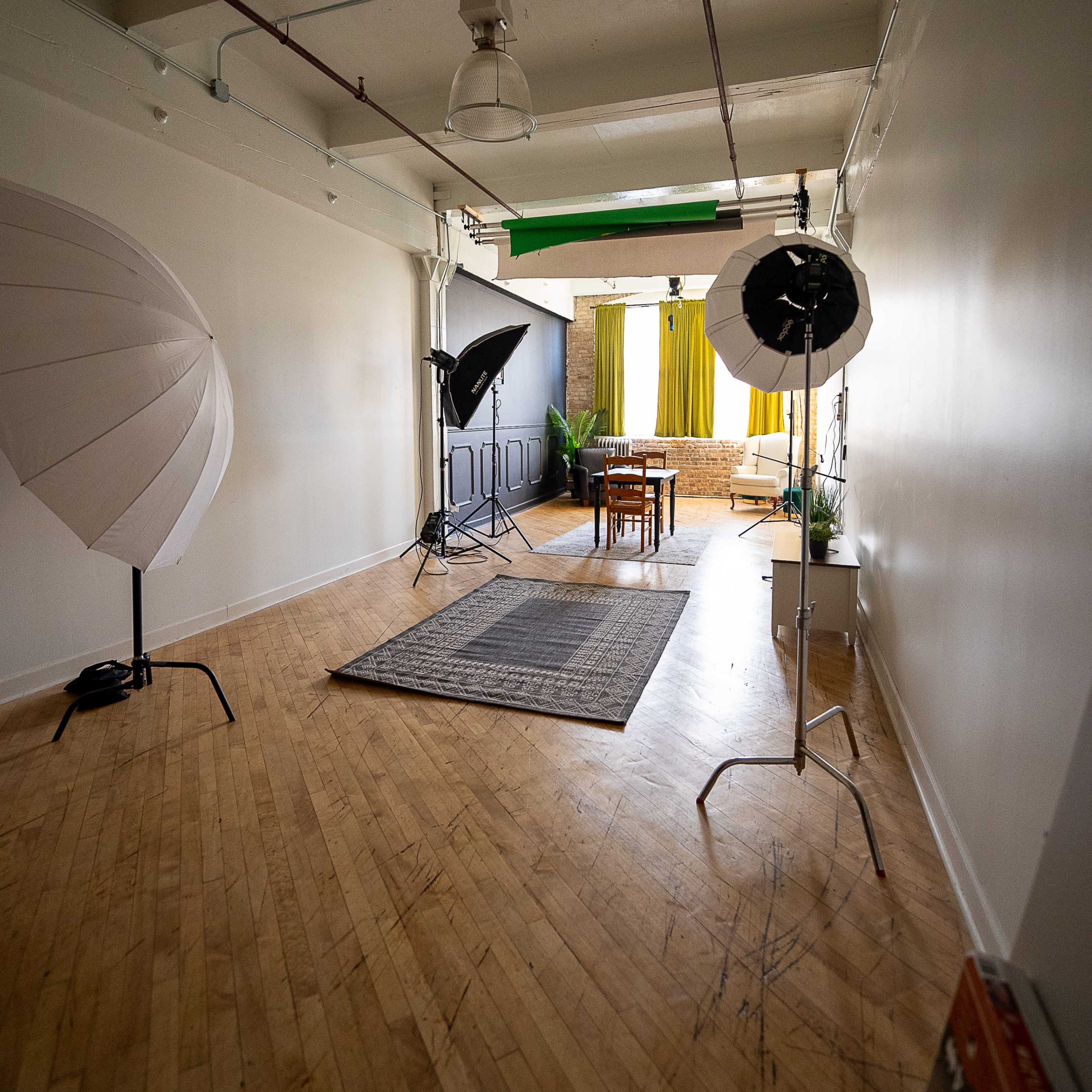 A photography studio with softboxes, a table, chairs, and a green backdrop against a wall.