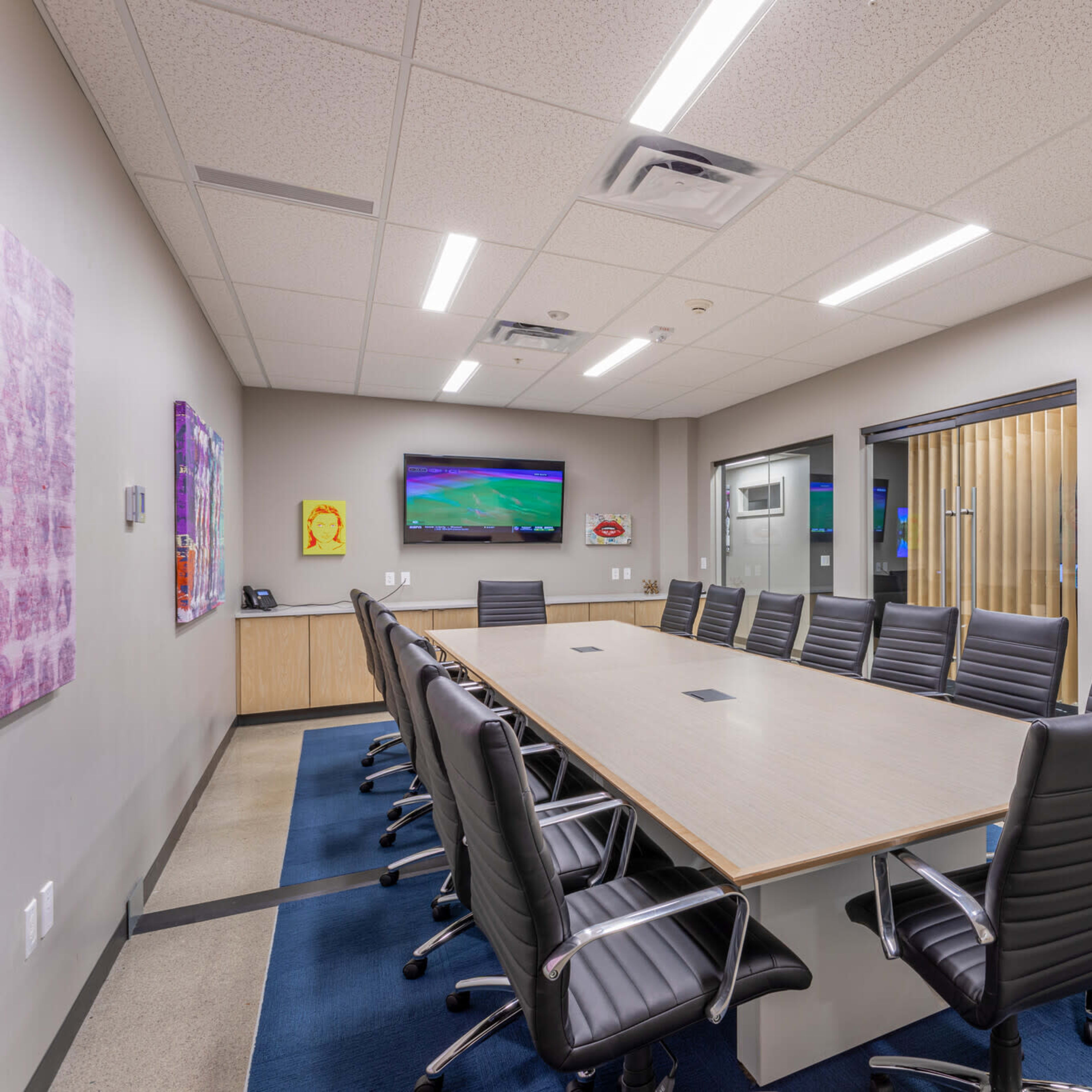 A modern conference room with a long rectangular table surrounded by black office chairs and a glass wall separating it from an adjacent room.